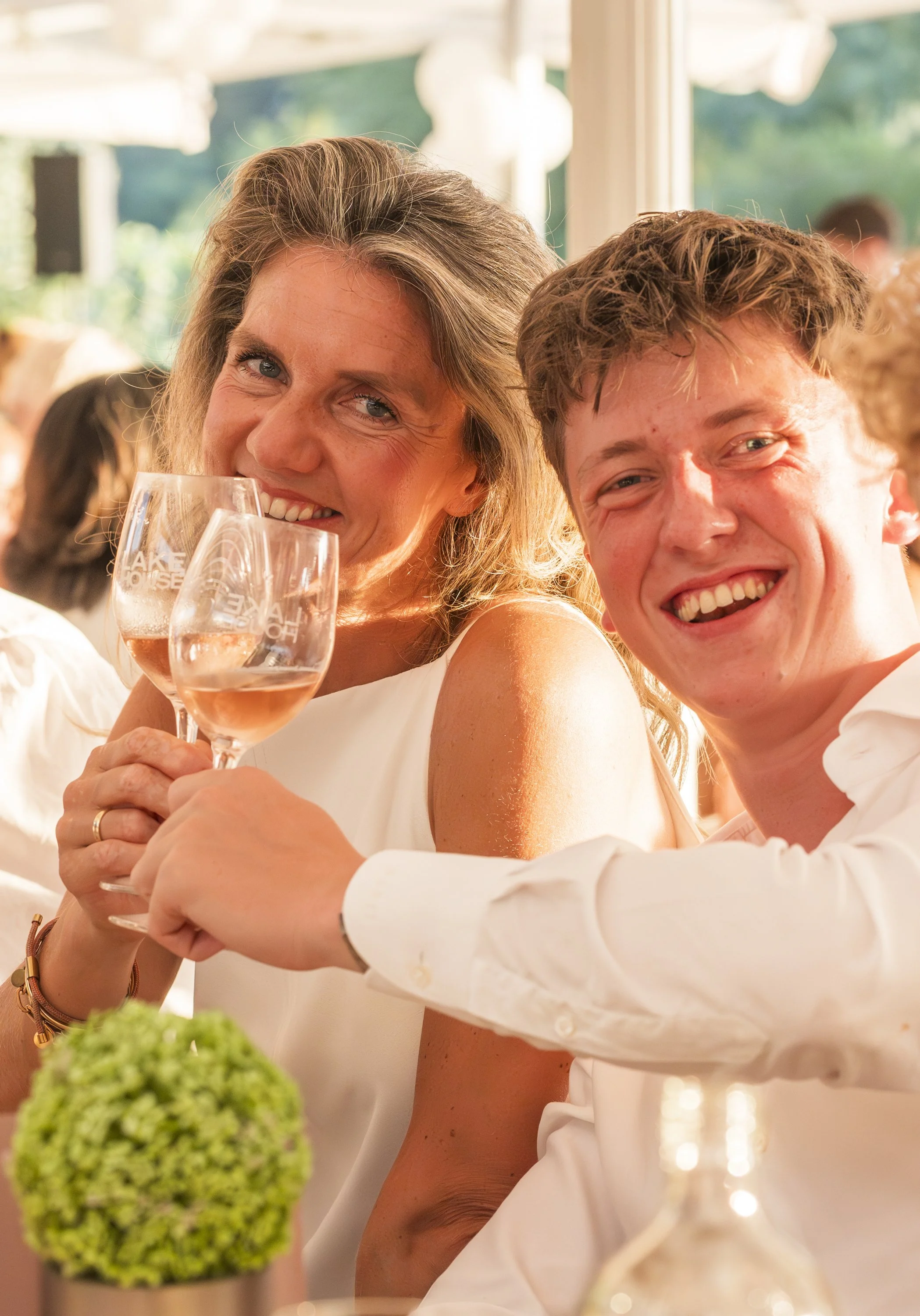 Twee lachende mensen tijdens een feest of bruiloft, een vrouw met een wijnglas en een man die naast haar zit, met bloemen en decoraties op de tafel.