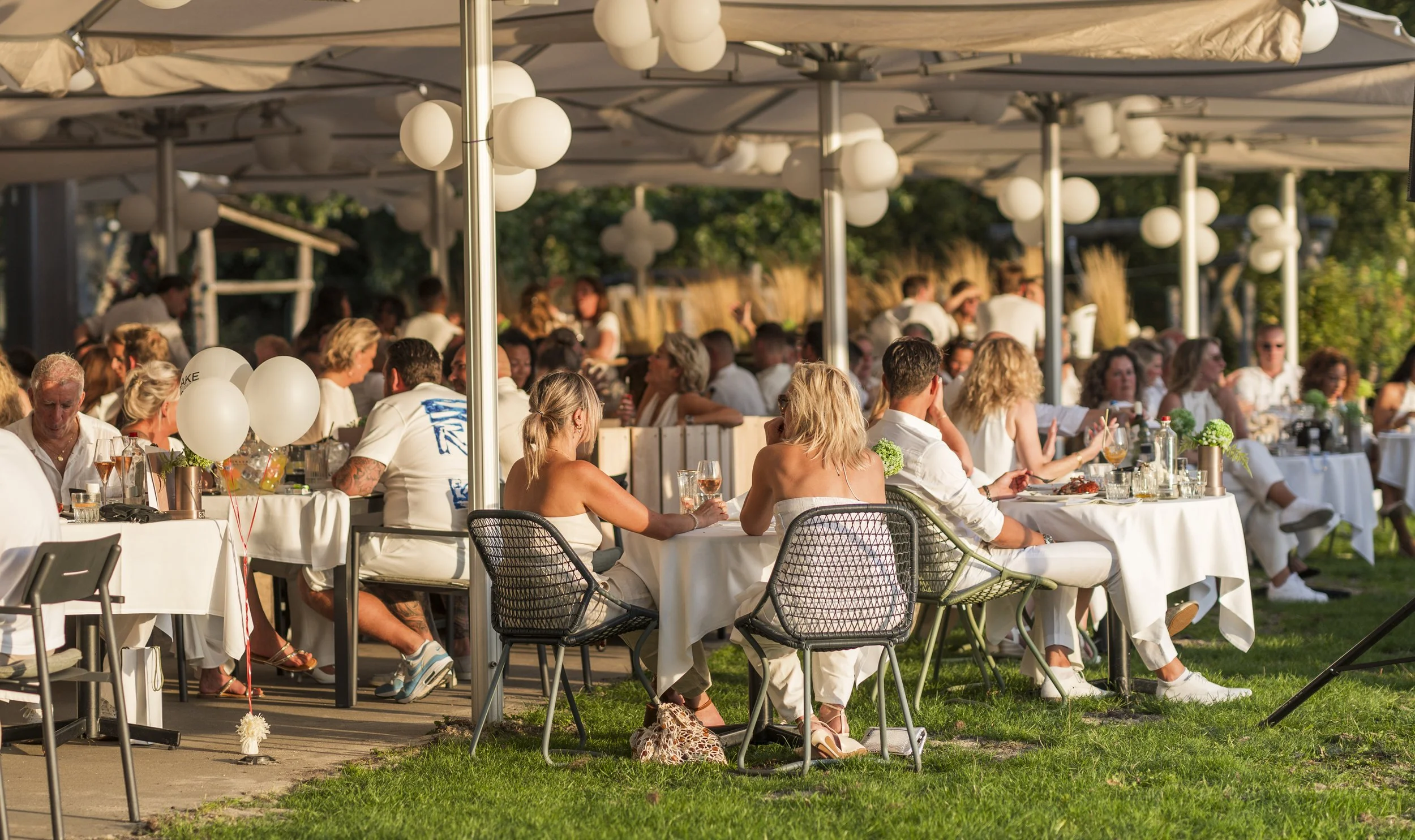 Gezellig buiten dinerscene met mensen op witte tafels met drankjes, omgeven door parasols en groene bomen in de avond.