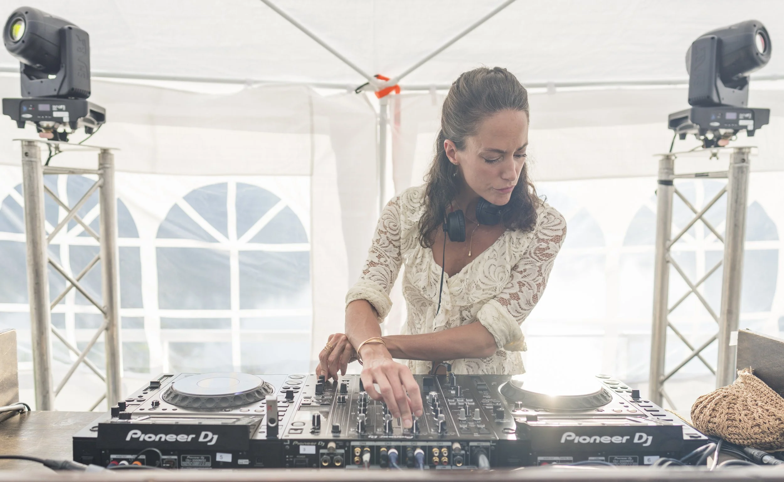 Vrouw met langen bruin haar en een witte kanten blouse achter een DJ-set, die zich voorbereidt om muziek te draaien. Ze zit onder een witte tent met grote ramen en omringd door professionele DJ- en lichtapparatuur.