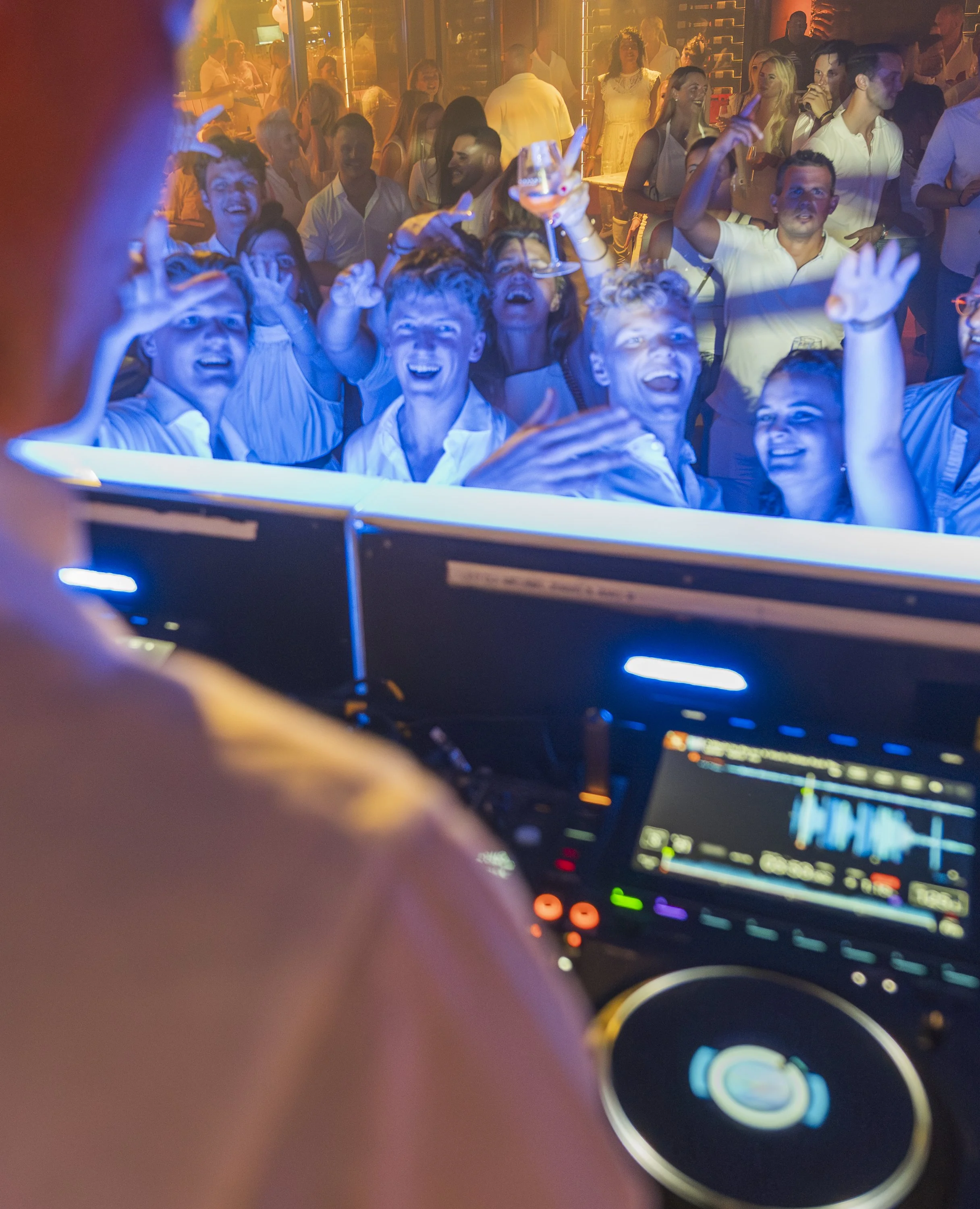 Beeld van een feestscène met mensen die dansen en lachen, gezien vanuit de dj-plek. De foto toont een menigte met witte kleding en een sfeer van plezier, met kleurrijke verlichting.