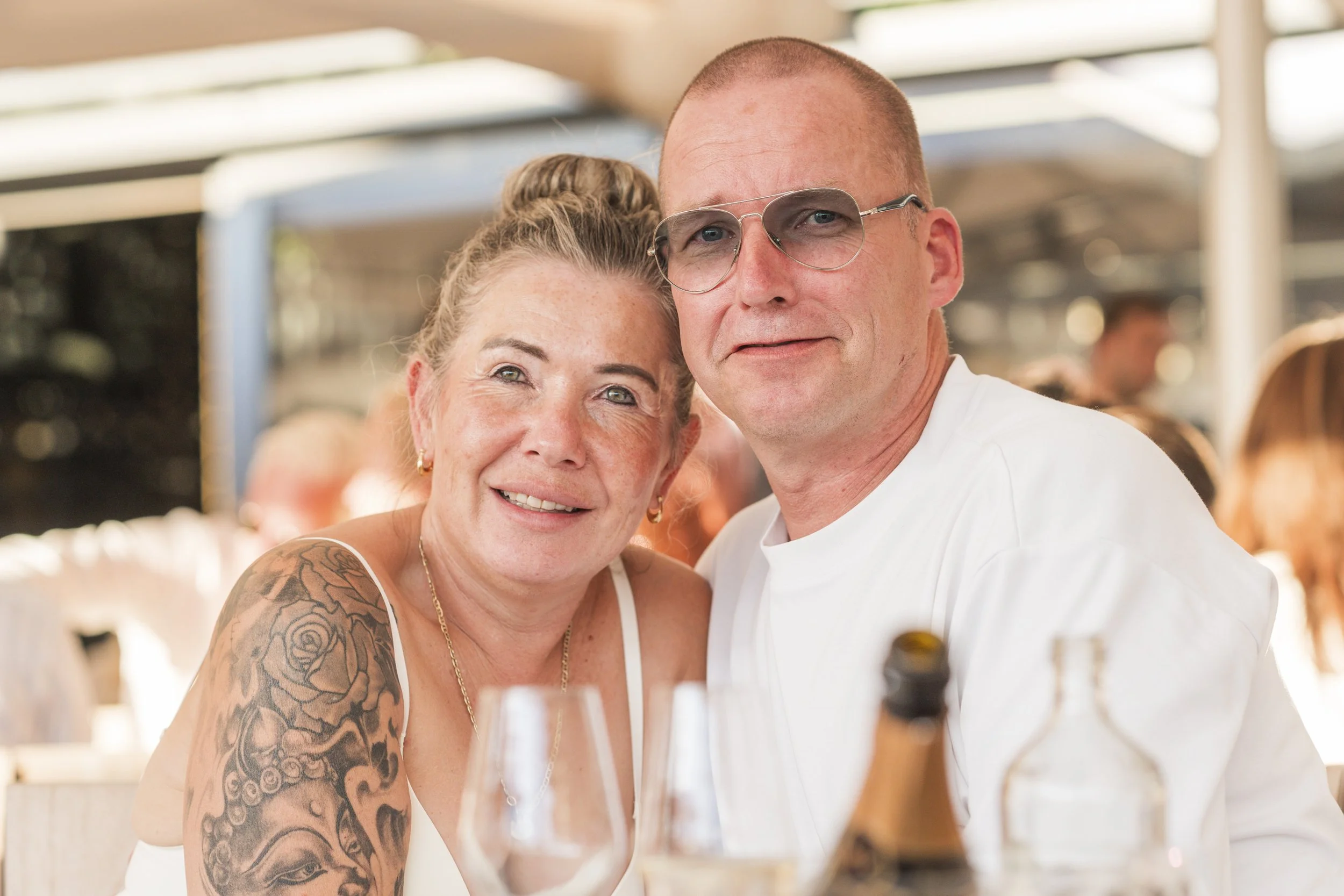 Een lachend oudere vrouw met tatoeages op haar arm en een jongere man zitten samen aan tafel tijdens een gelegenheid, mogelijk een bruiloft of familiefeest.