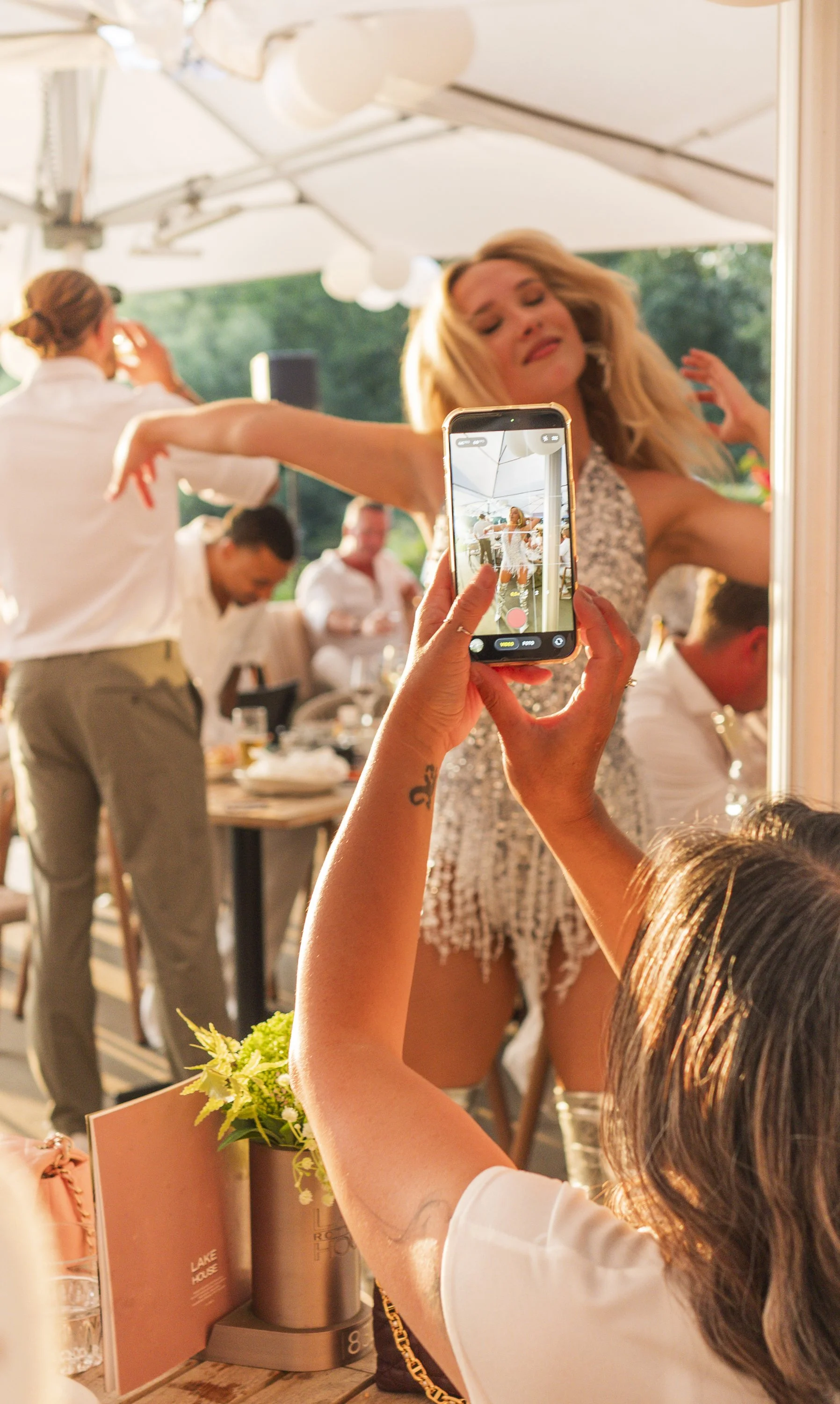 Een vrouw maakt foto's van een danseres tijdens een feest of evenement dat buiten plaatsvindt. De danseres is aan het dansen en er zitten meerdere mensen aan tafel op de achtergrond.