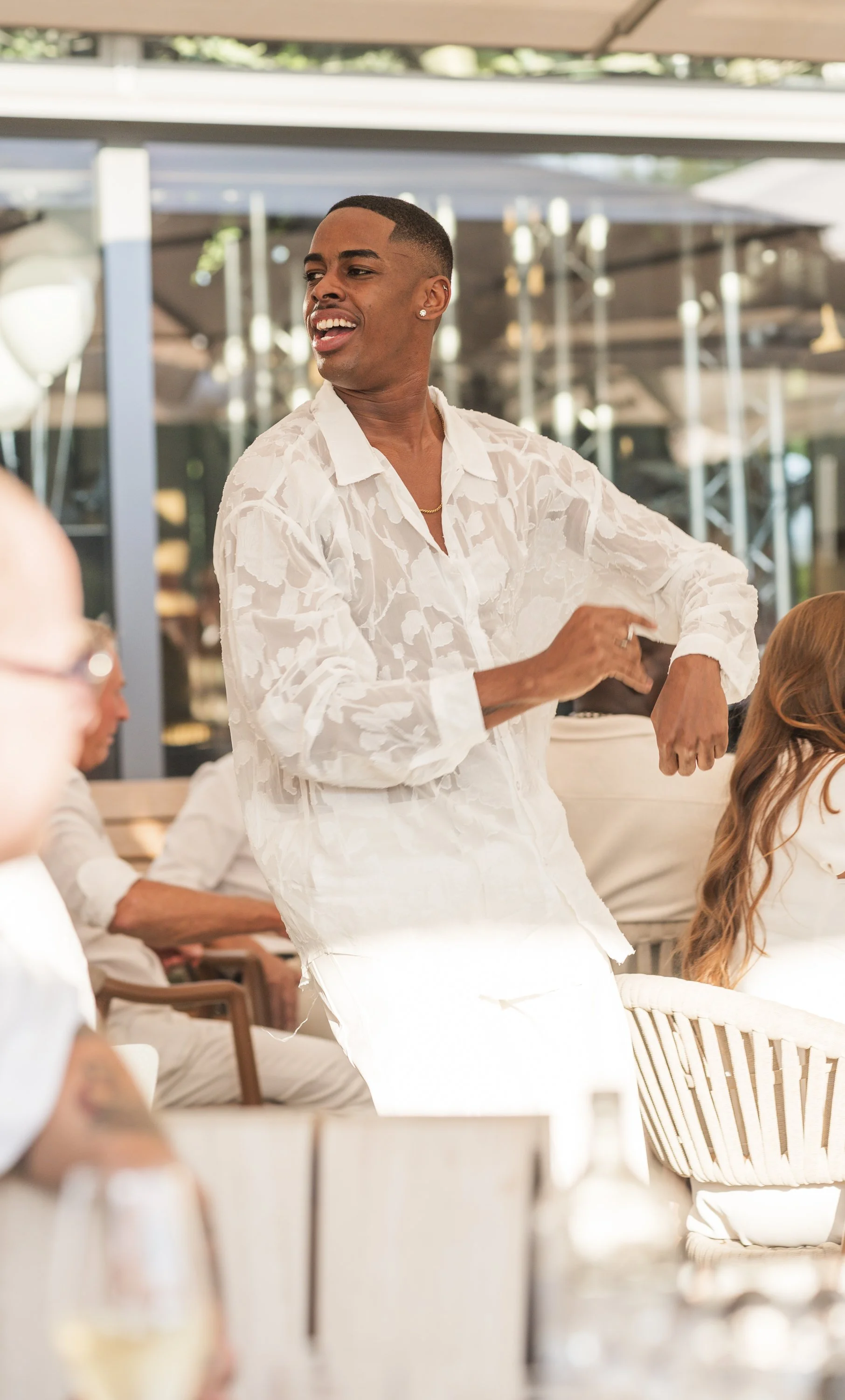 Een persoon met moderner kapsel en een witte transparante blouse, die lijkt te dansen of bewegen in een lichte, moderne ruimte, met mensen die aan tafel zitten.