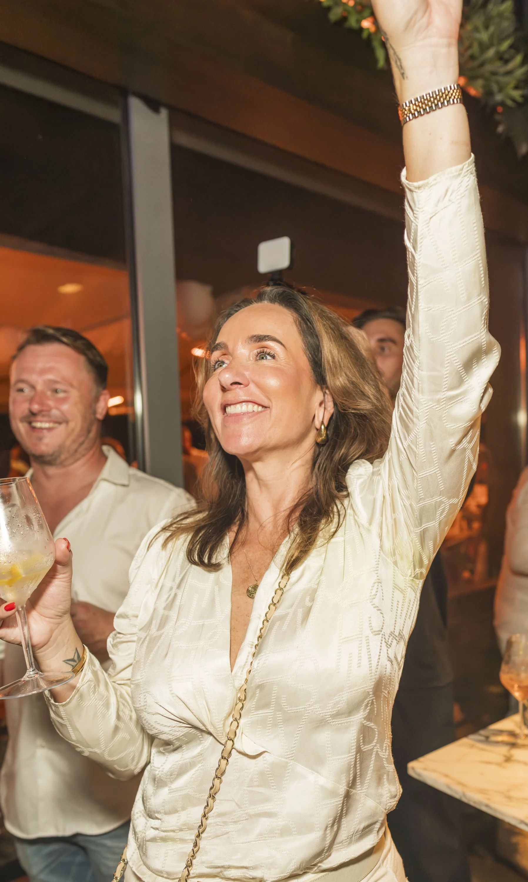 Een vrouw met bruin haar die lacht en haar hand omhoog steekt tijdens een feestje in een interieur met warme verlichting, terwijl ze een glas wijn vasthoudt.