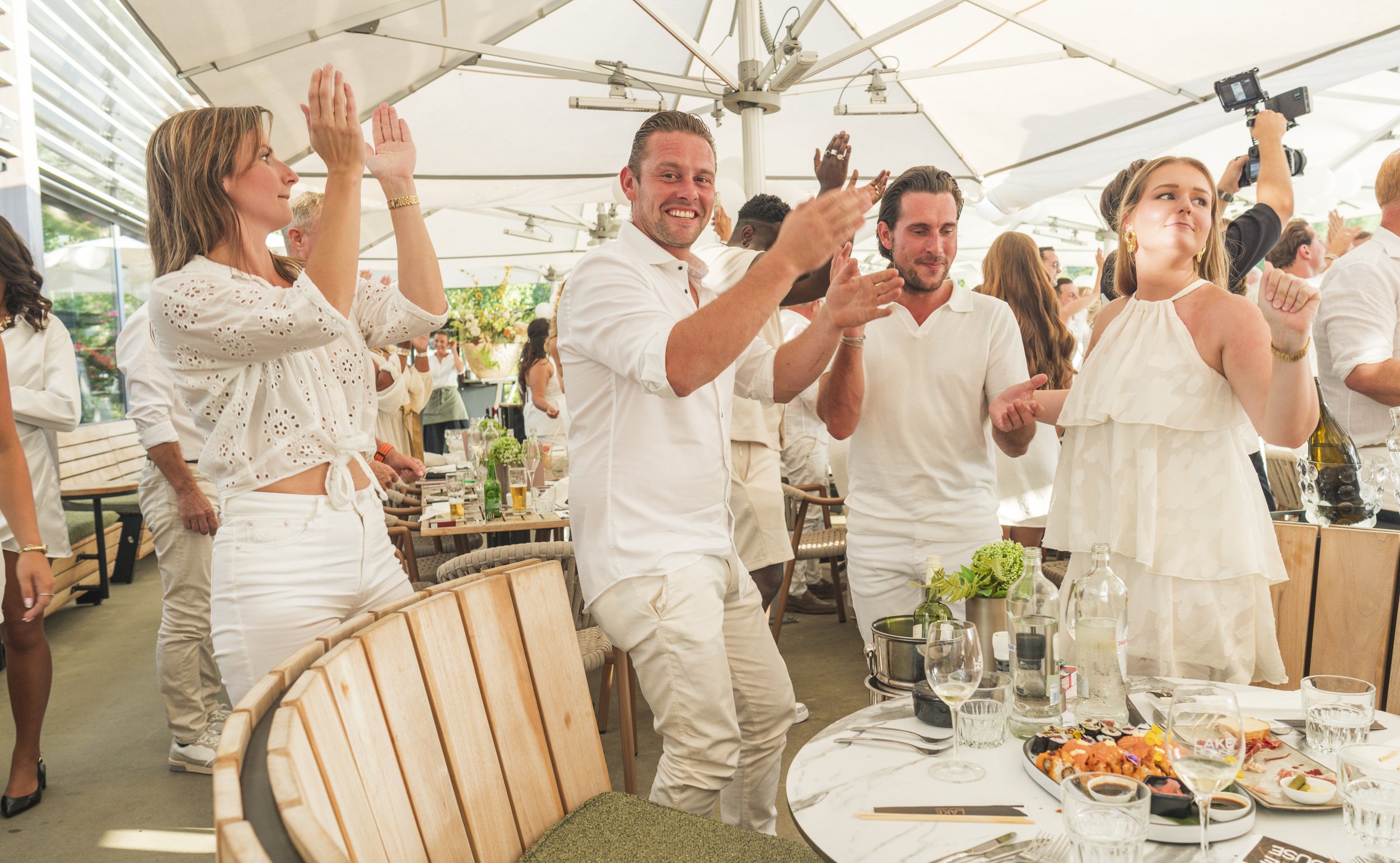 Mensen vieren feest in een restaurant onder een grote parasol, ze staan en applaudisseren terwijl ze genieten van eten en drinken.