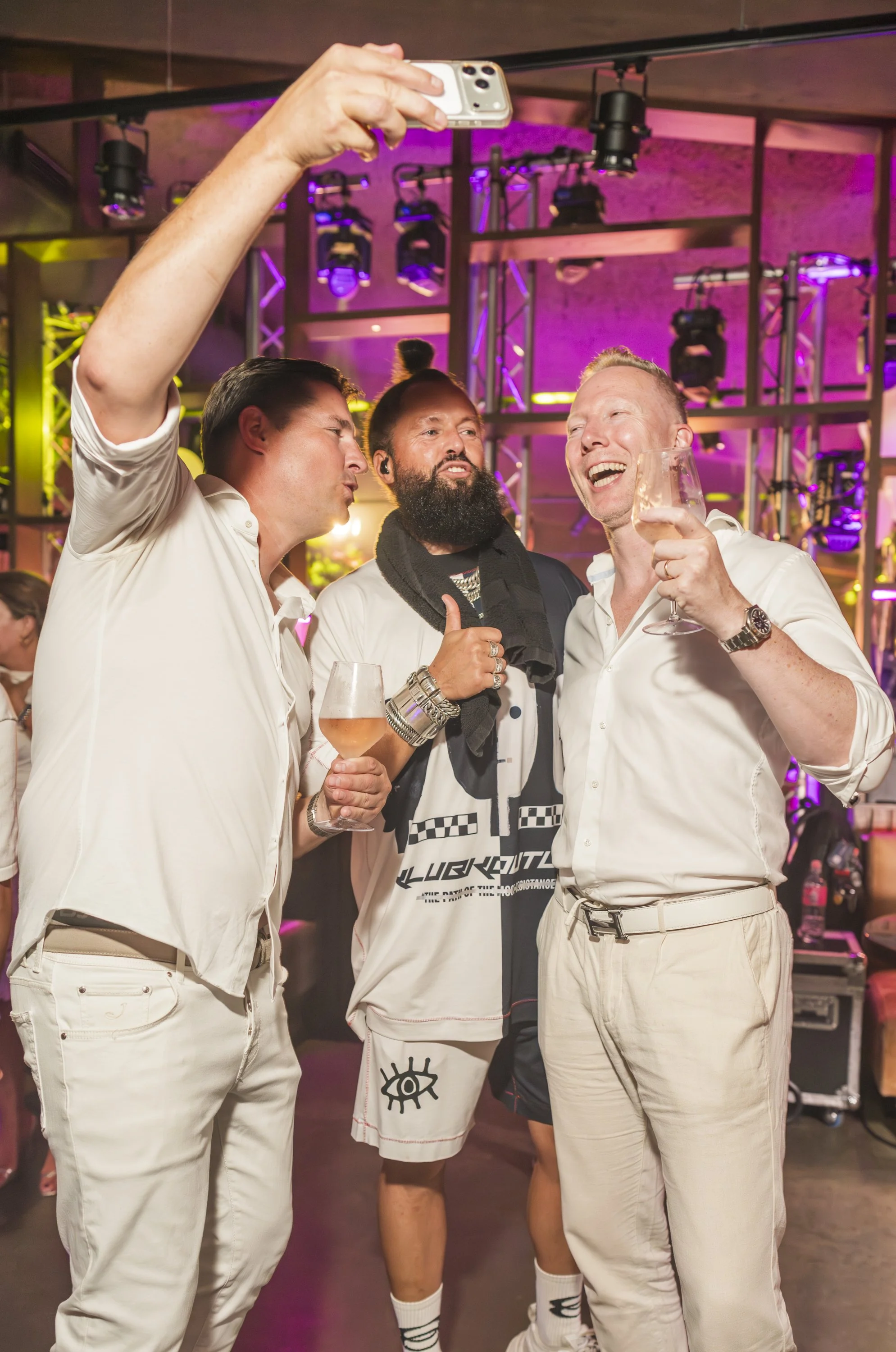 Drie mannen maken een selfie in een feestlocatie met kleurrijk licht, één man houdt een glas rosé-wijn vast en onzeidt naar de camera.