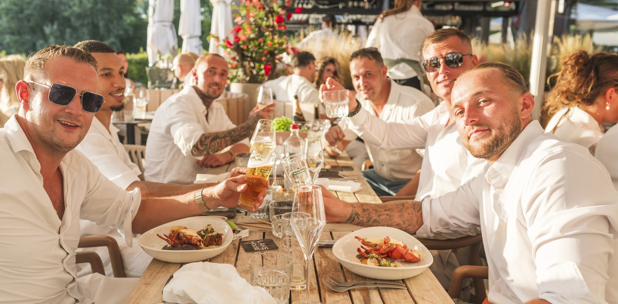 Een groep mannen zit aan een lange tafel tijdens een buitenmaaltijd, met eten en drinkglazen voor zich, genietend van een zonnige dag.