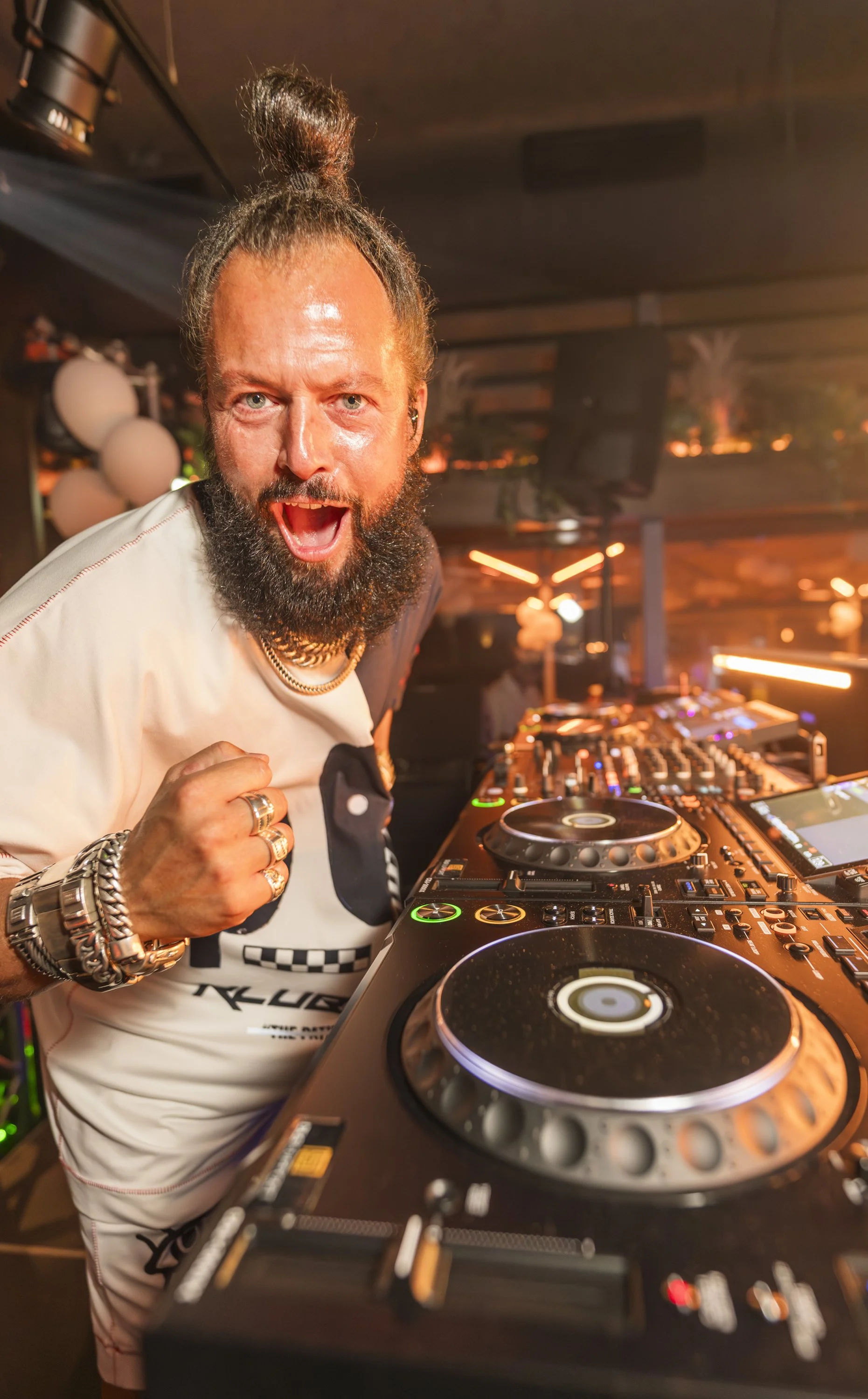 Man met een ponytail, baard en sieraden, die DJ apparaten bedient in een club. Hij heeft een opwindende uitdrukking.