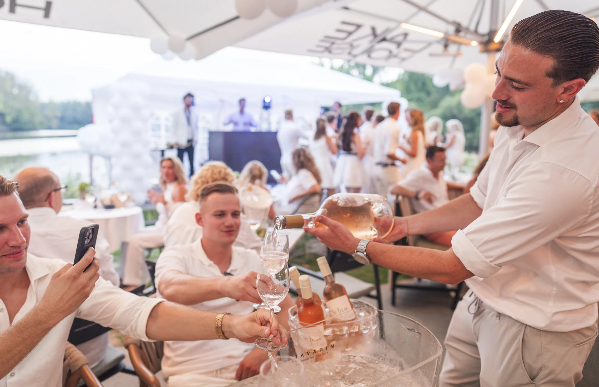 Man serveert een glas roséwijn aan een man die een smartphone vasthoudt tijdens een feest met veel mensen onder een witte tent.