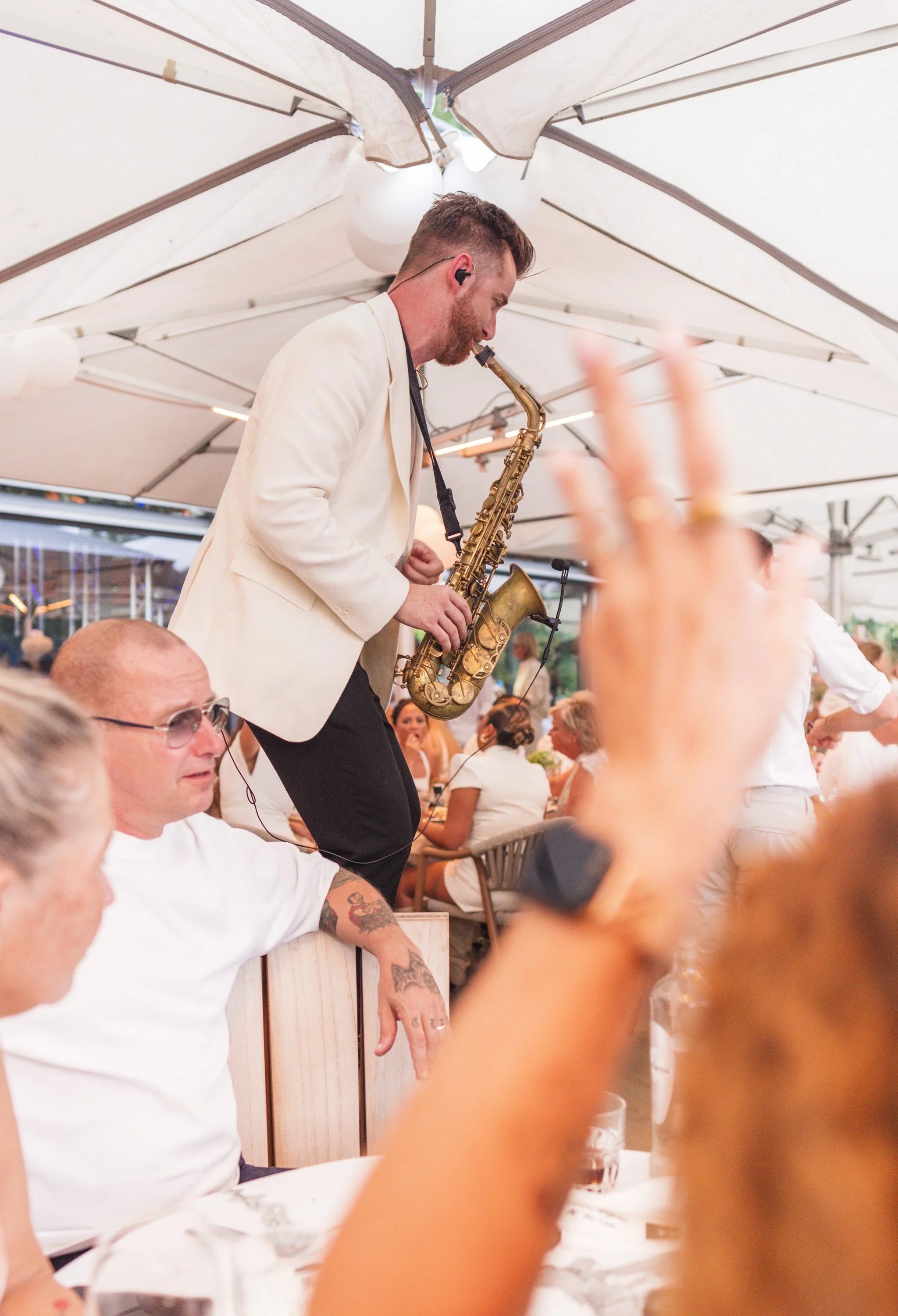 Een man met een baard en een witte jas speelt saxofonist tijdens een liveoptreden onder een grote parasol. De sfeer lijkt levendig met mensen die genieten en een feestelijke ambiance.