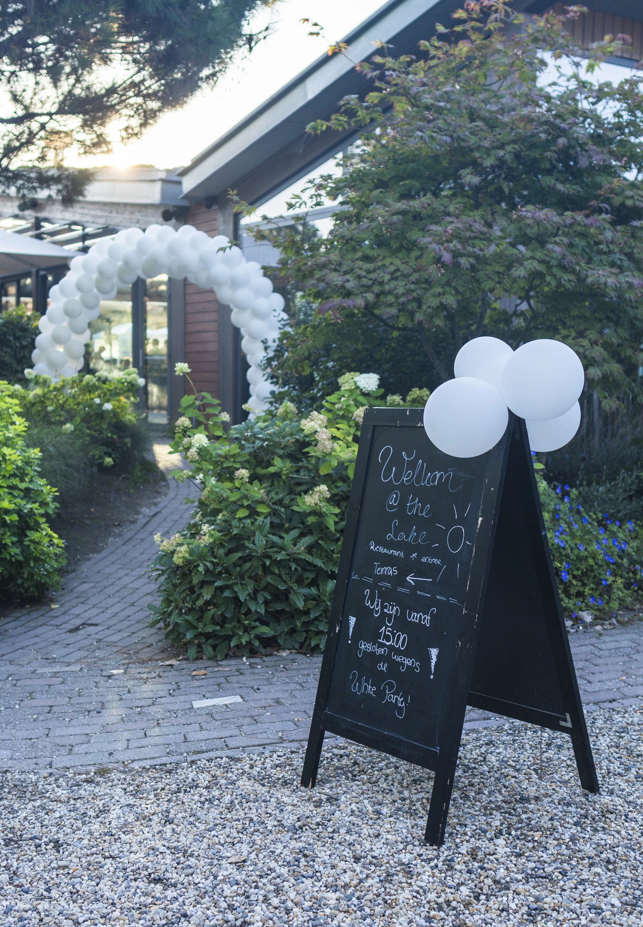 Een zwart uithangbord met witte tekst en een hartvormige witte ballon, geplaatst op een stenen oprit met groene planten en bloemen. In de achtergrond is een tuin met een boog van witte ballonnen en een gebouw te zien, met de zon die ondergaat.