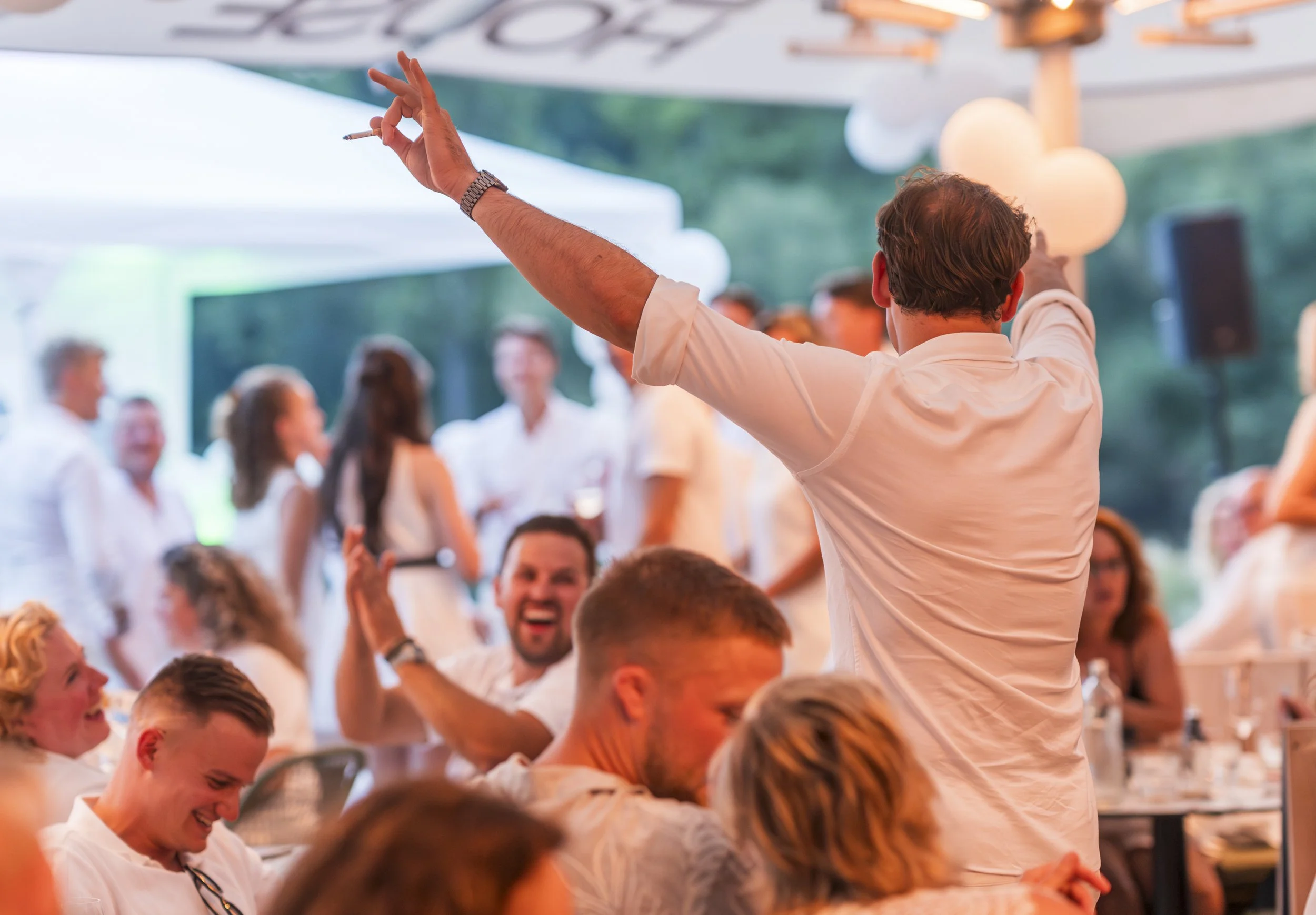 Man met bruin haar en witte shirt die uitdrukkelijk zwaaien met een hand die een sigaret vasthoudt, terwijl hij op een feest of bijeenkomst staat met een menigte mensen op de achtergrond onder een witte tent of overkapping.