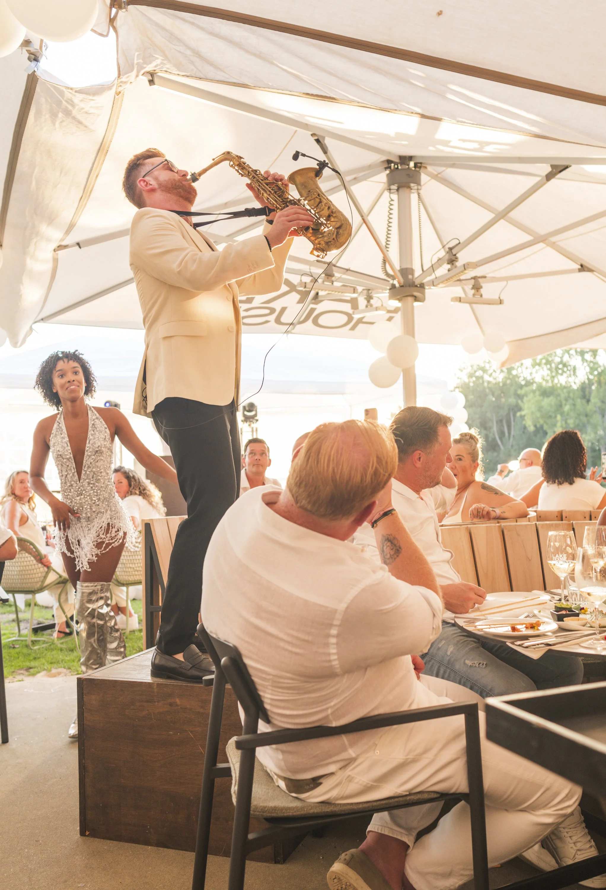 Muzikant die op een houten podium een elektrische saxofoon speelt onder een grote witte parasol, terwijl een man in een wit T-shirt in een restaurant zit en naar de performer kijkt, omgeven door andere eters en een vrouwelijke danser met haltertop en