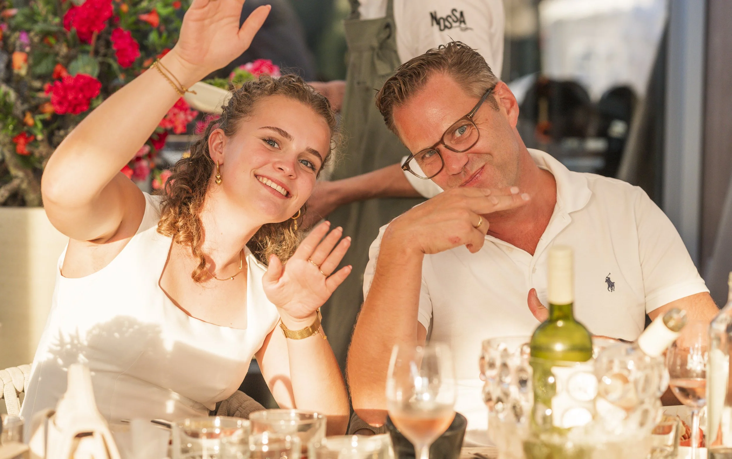 Een vrouw en een man zitten aan een tafel in een restaurant, de vrouw zwaait naar de camera en lacht, de man draagt een bril en wijst naar zichzelf, er zijn wijn en eten op de tafel, bloemen op de achtergrond, de sfeer is vrolijk en gezellig.