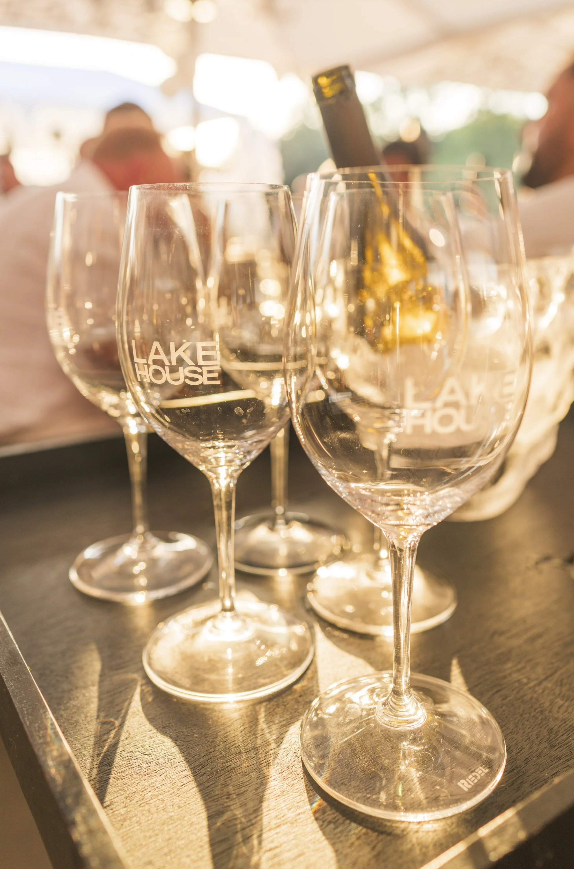 Glazen met 'LAKE HOUSE' logo en een fles wijn op een tafel tijdens een evenement of bijeenkomst.