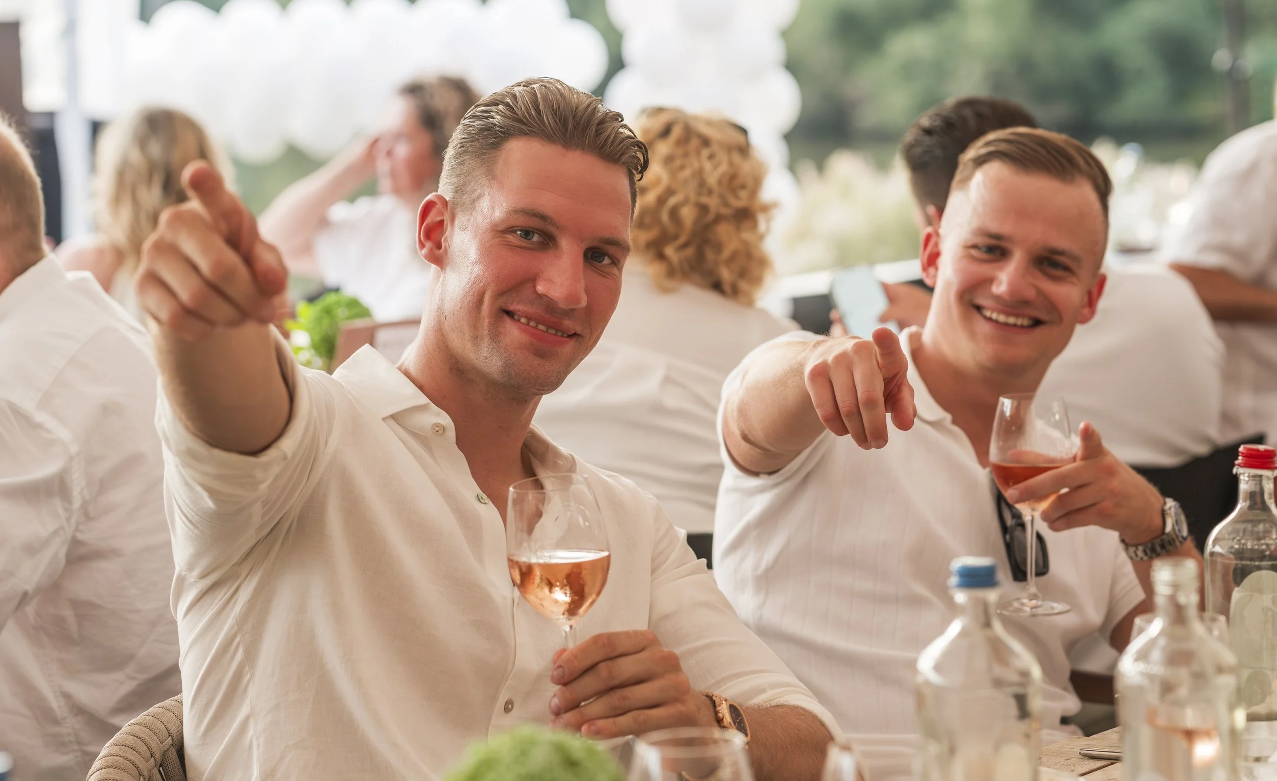 Vrienden genieten van een sociale bijeenkomst, proosten met rosé wijn, zittend aan een tafel met drankjes en snacks, buiten met groene achtergrond.