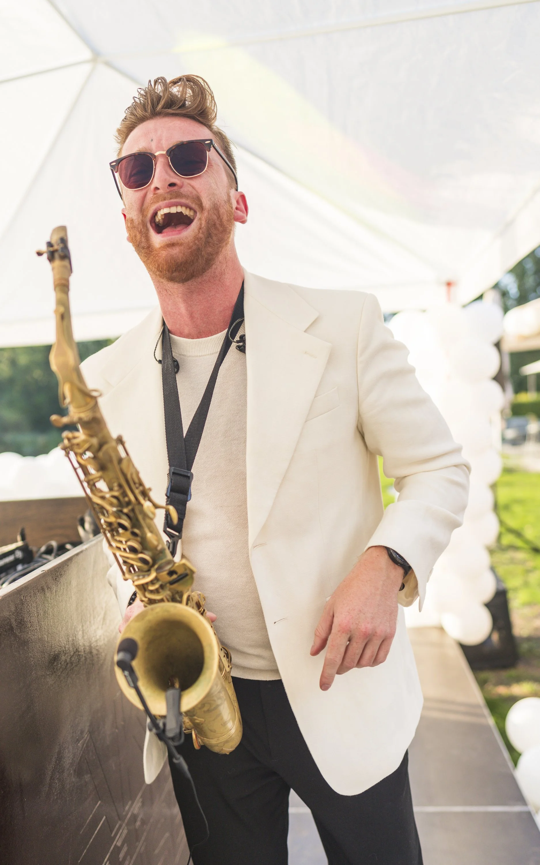 Man met rode baard en sunglasses die een saxofon vasthoudt op een feest met witte ballonnen onder een tentdak.
