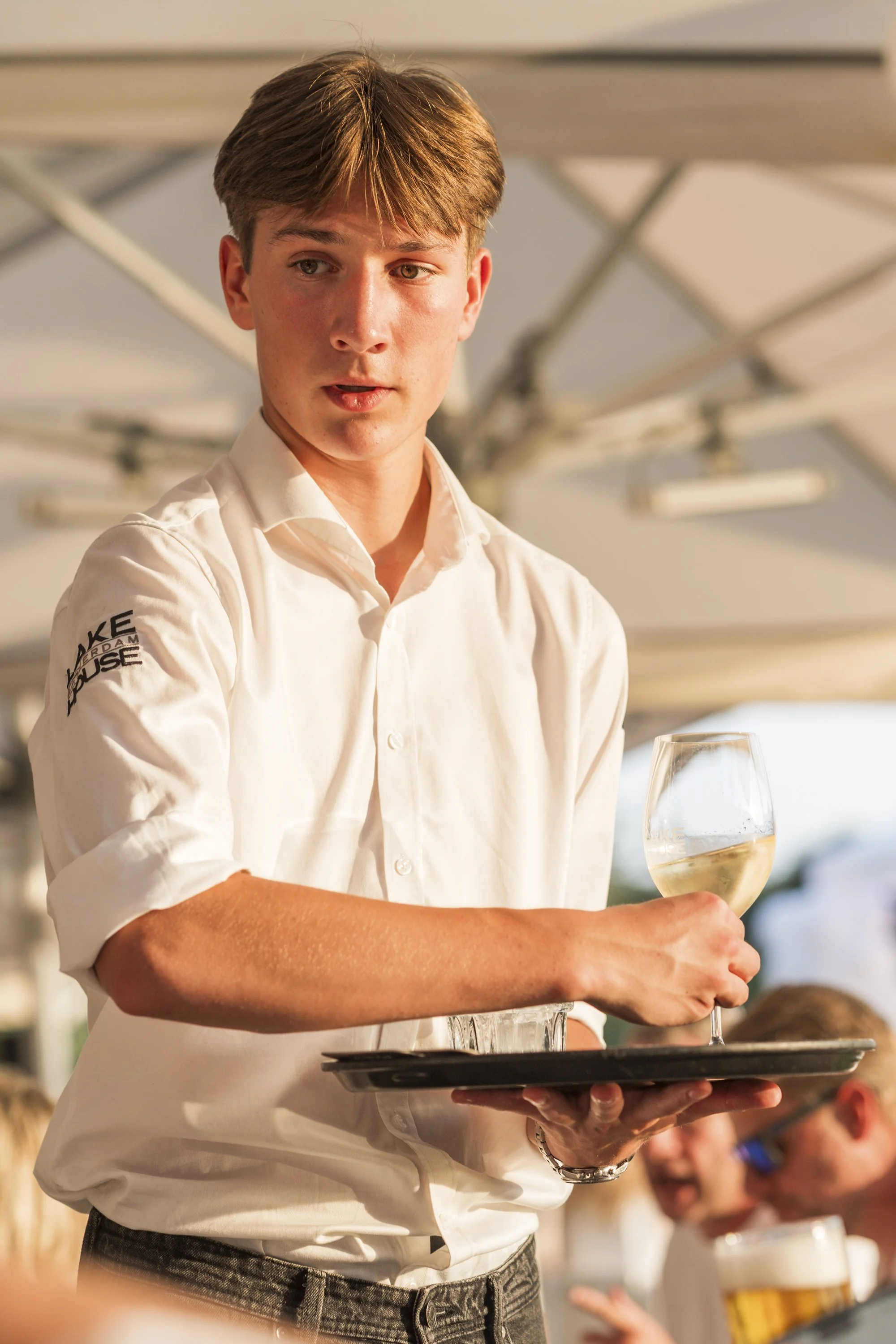 Een jonge ober in een witte overhemd serveert een glas witte wijn op een dienblad in een restaurant.