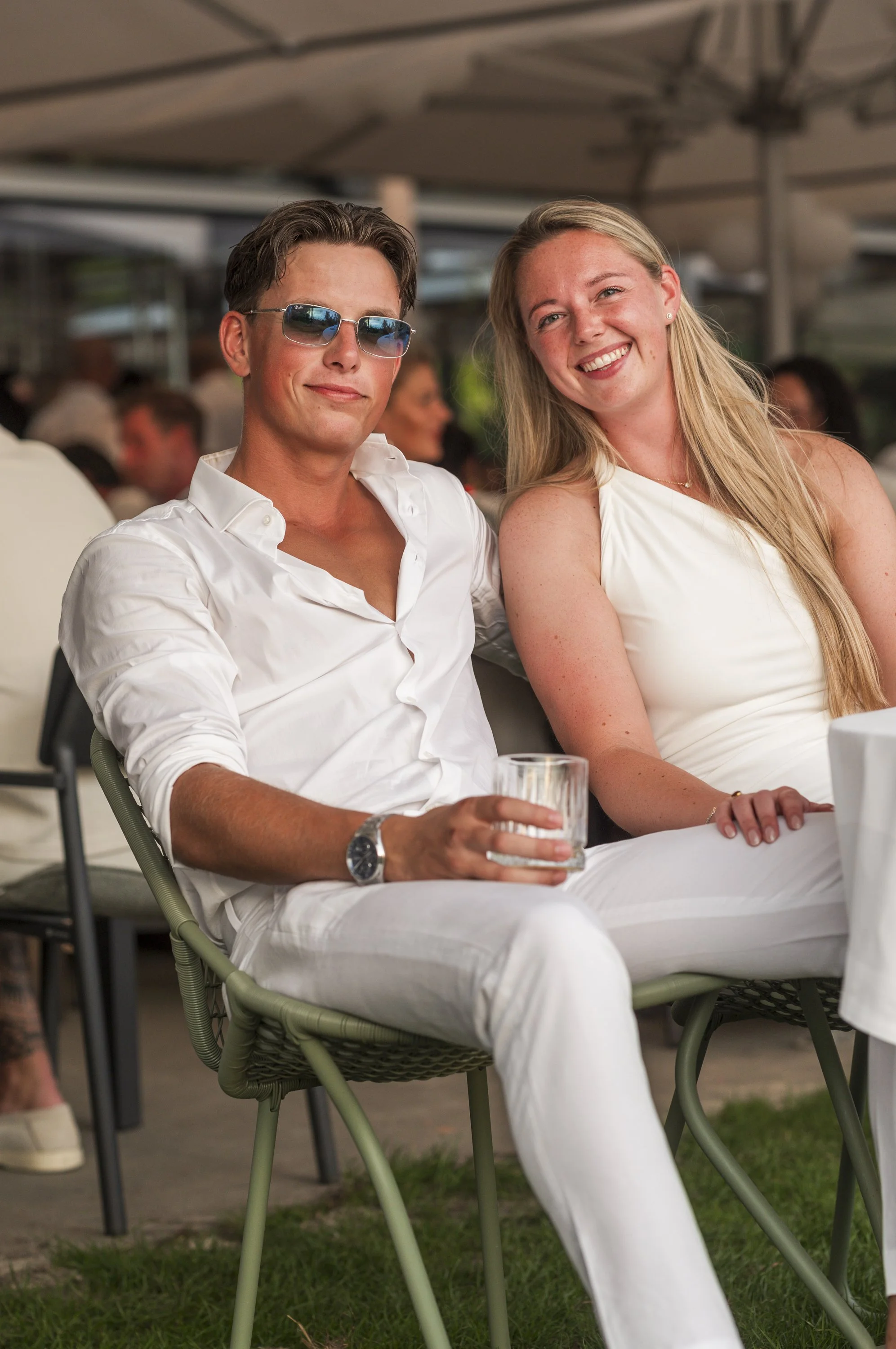 Twee mensen zitten buiten onder een parasol, de man draagt een witte overhemd en zonnebril, de vrouw draagt een witte jurk, hij houdt een glas water vast, beiden glimlachen.