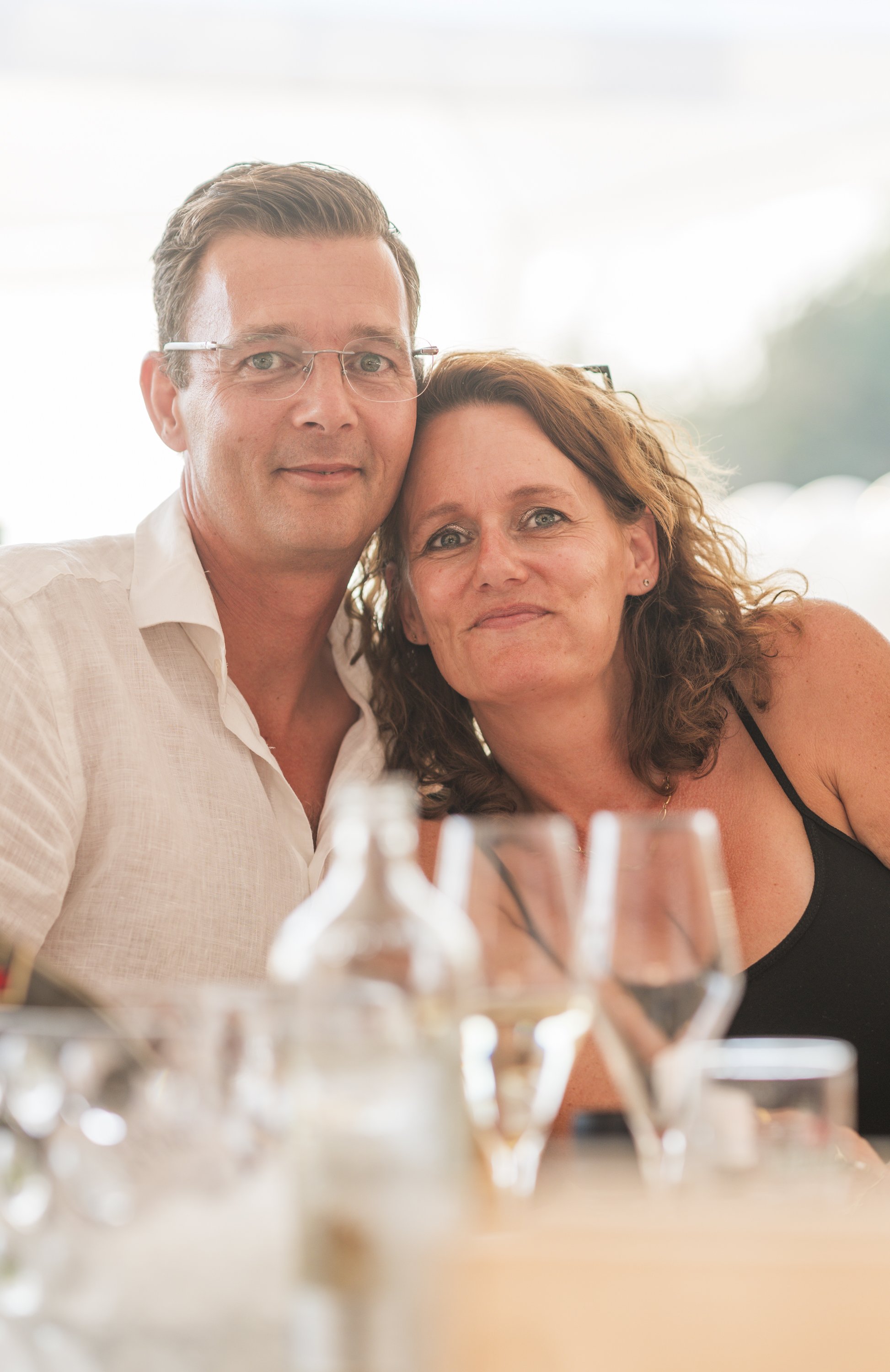 Een man met kort haar en een vrouw met krullend haar poseren dicht bij elkaar, ze zitten aan een tafel met enkele glazen en flesjes, waarschijnlijk tijdens een feest of diner.