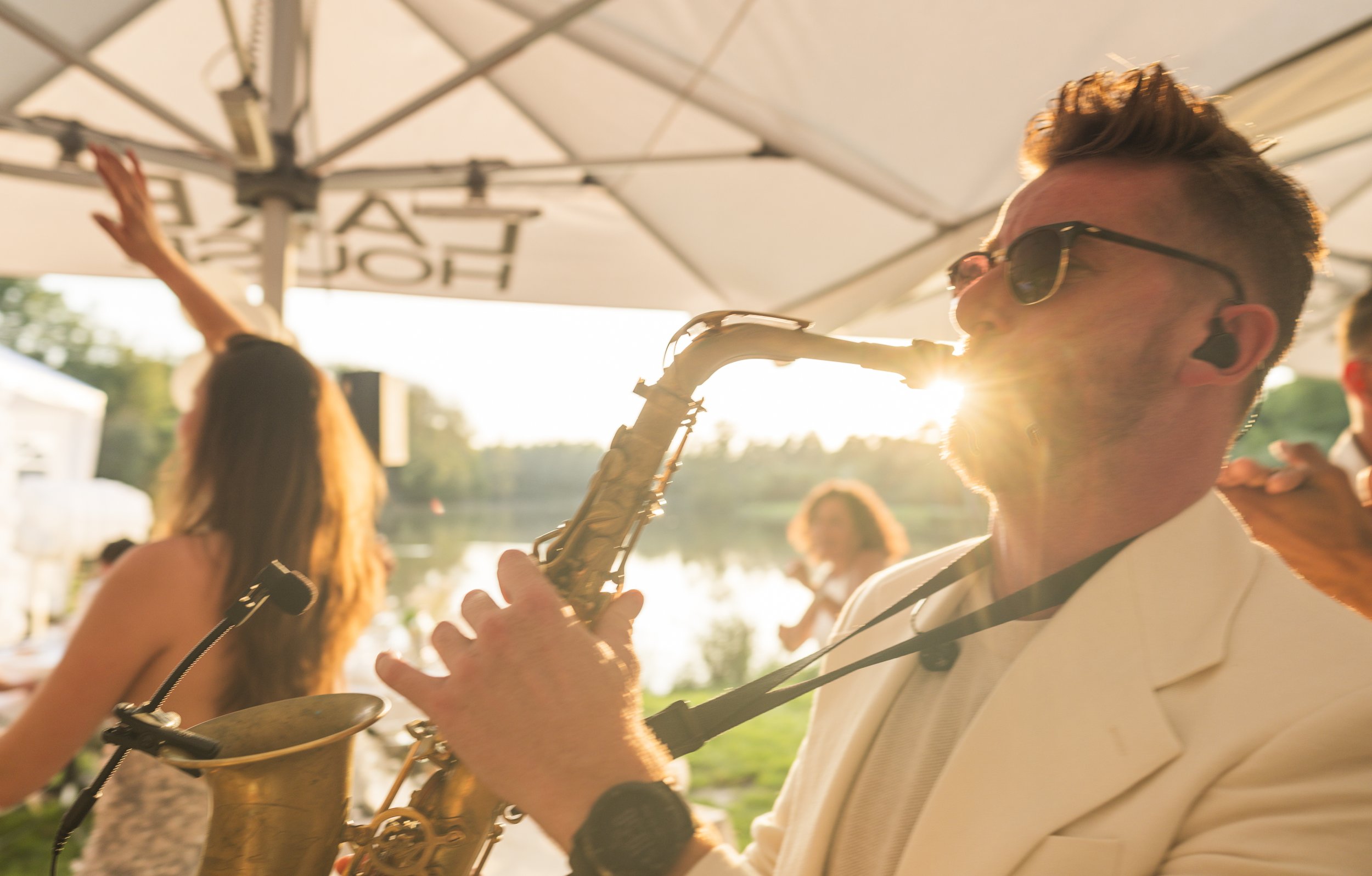 Man speelt saxofoon op een zonnig buitenevenement, met mensen op de achtergrond en een groene omgeving.