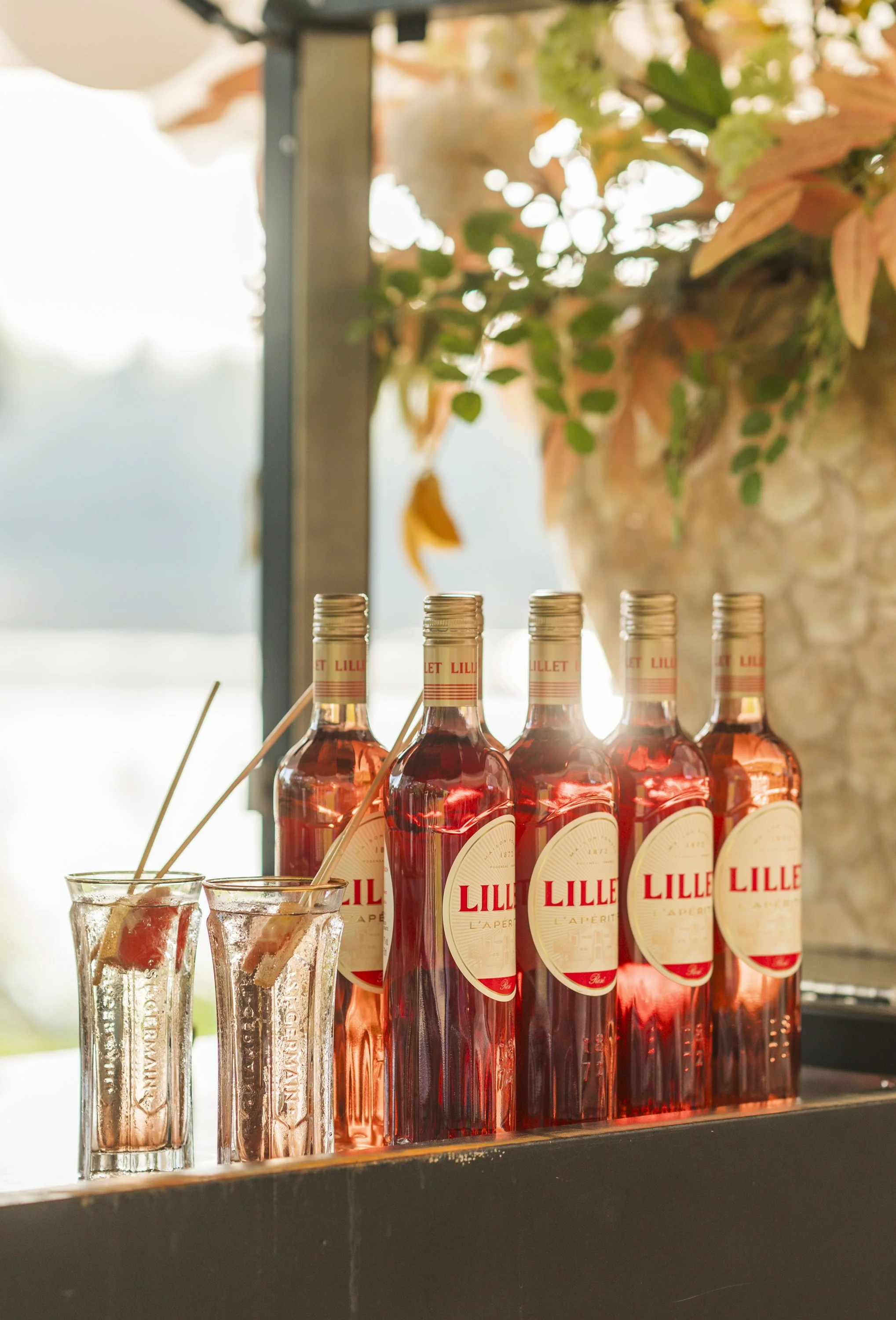 Verschillende flesjes rosé wijn met op de fles het label 'Lillet' staan, naast twee glazen met ijs en een roerstaafje, op een barkruk met een achtergrond van een grote plant en een raam met uitzicht op een lichten en buiten.