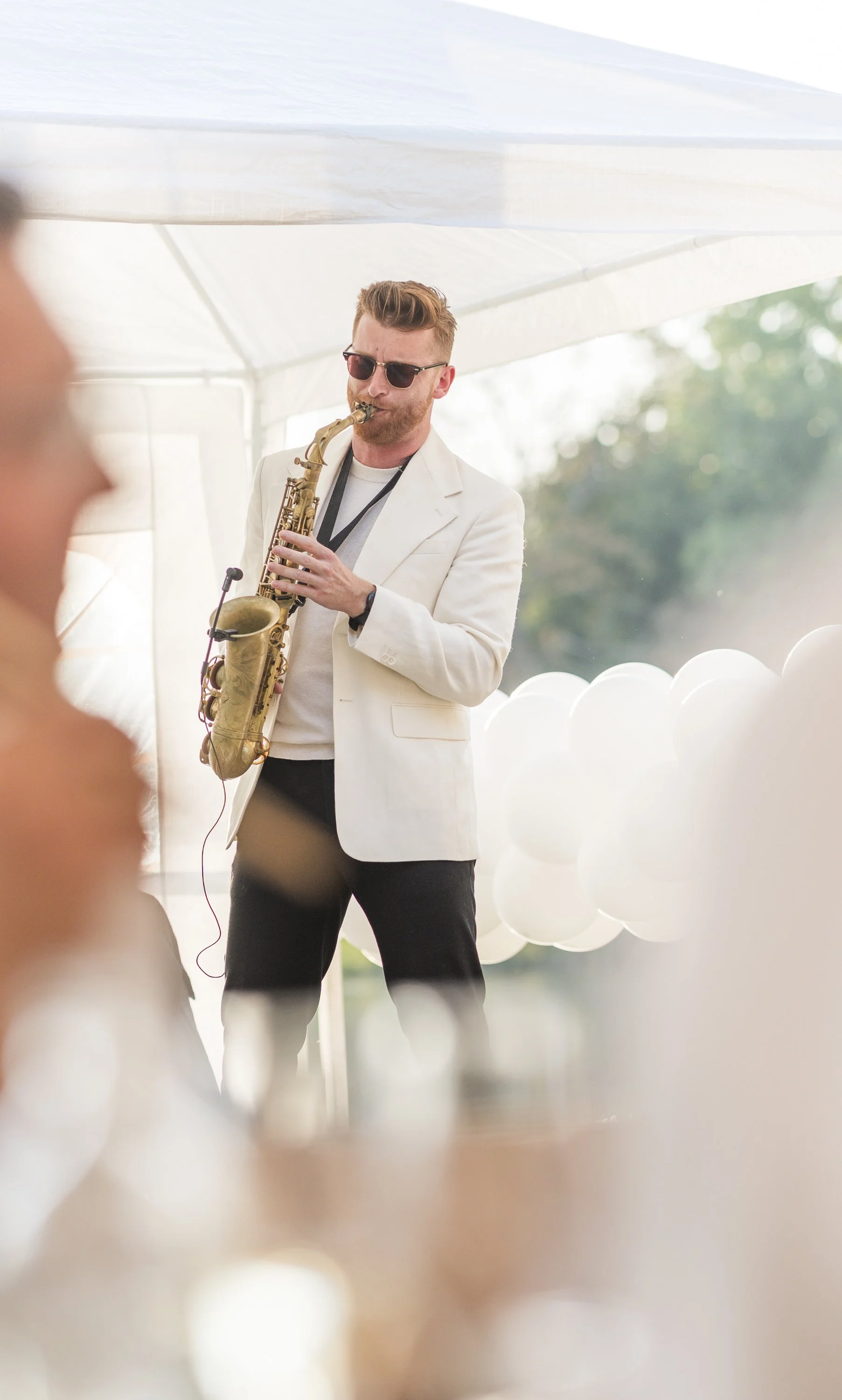 Man met een lichte jas en zonnebril speelt saxofoon op een buitengebeurtenis, omringd door witte ballonnen.