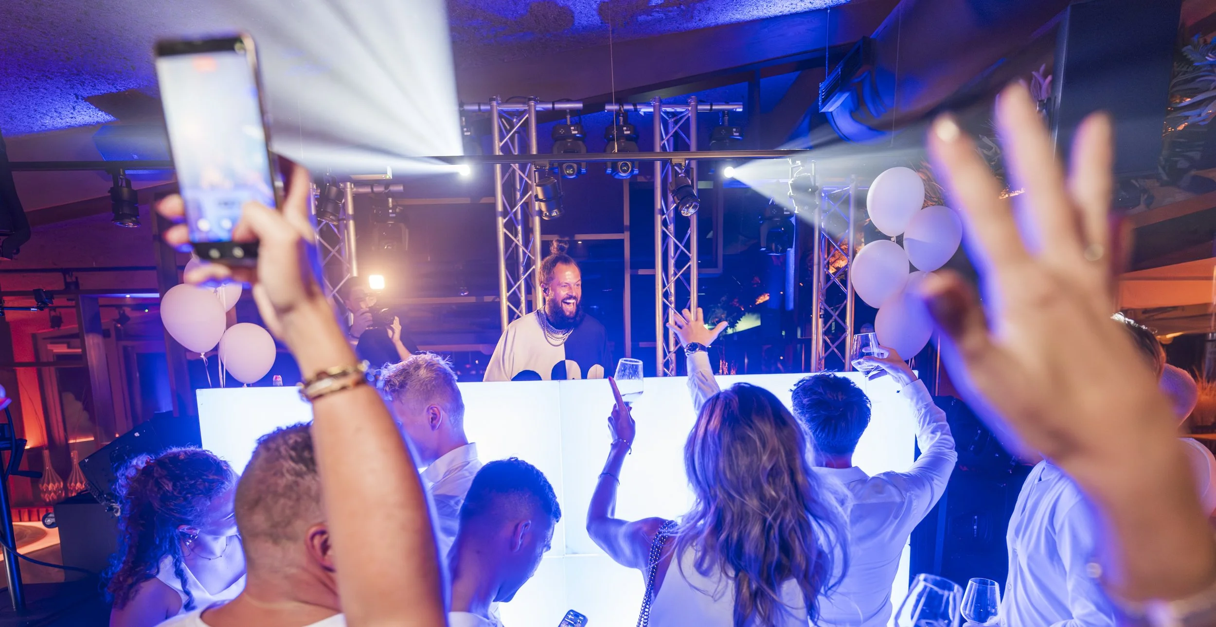 Mensen op een feestje dansen en genieten bij een DJ die achter een lichtbord staat, met ballonnen en kleurrijk verlichtingslicht op de achtergrond.
