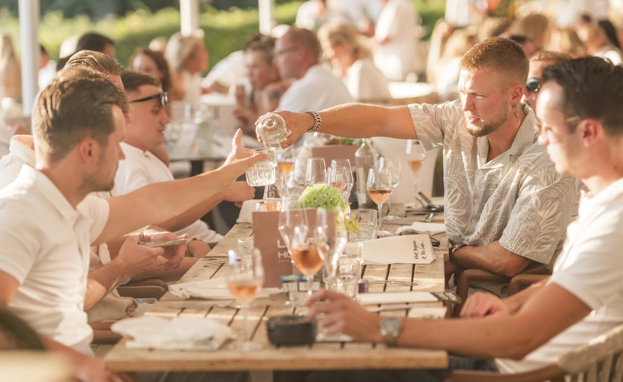 Een groep mensen zit samen aan een lange houten tafel buitenshuis onder een overkapping. Een man schenkt drinken in een glas van een andere persoon aan de tafel. De sfeer lijkt ontspannen en feestelijk met veel mensen die praten en lachen.