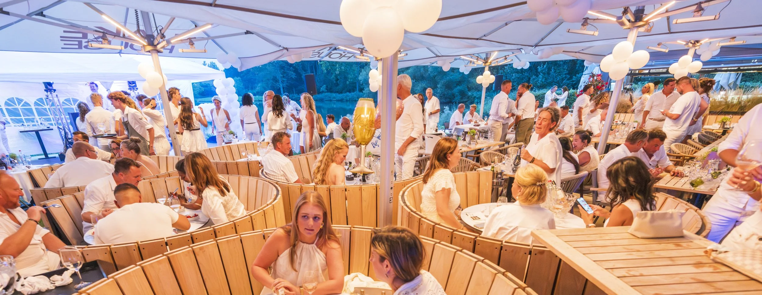 Een zomerse avondfeest aan het water met mensen in witte kleding onder grote parasols versierd met witte ballonnen.