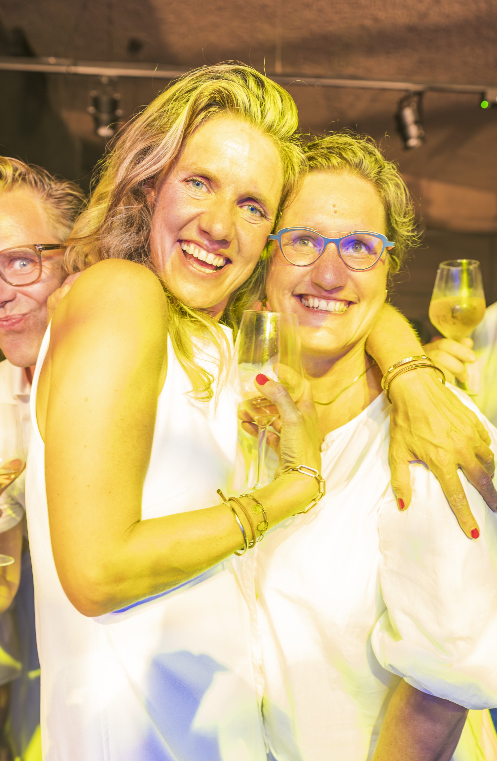 Vrouw die glimlacht en een glas witte wijn vasthoudt, omringd door anderen, vieren feest in een binnenruimte.