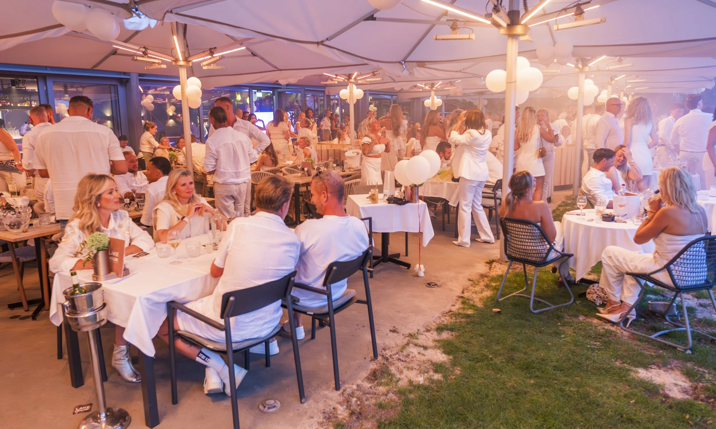 Een feestelijke bijeenkomst met mensen die binnen en buiten een versierd ruim bijkaarten, rijst, en tafels met witte tafelbekleding. Gasten zitten, staan en praten, terwijl ballonversieringen en grote parasols de ruimte decoreren. Mensen dragen voorn