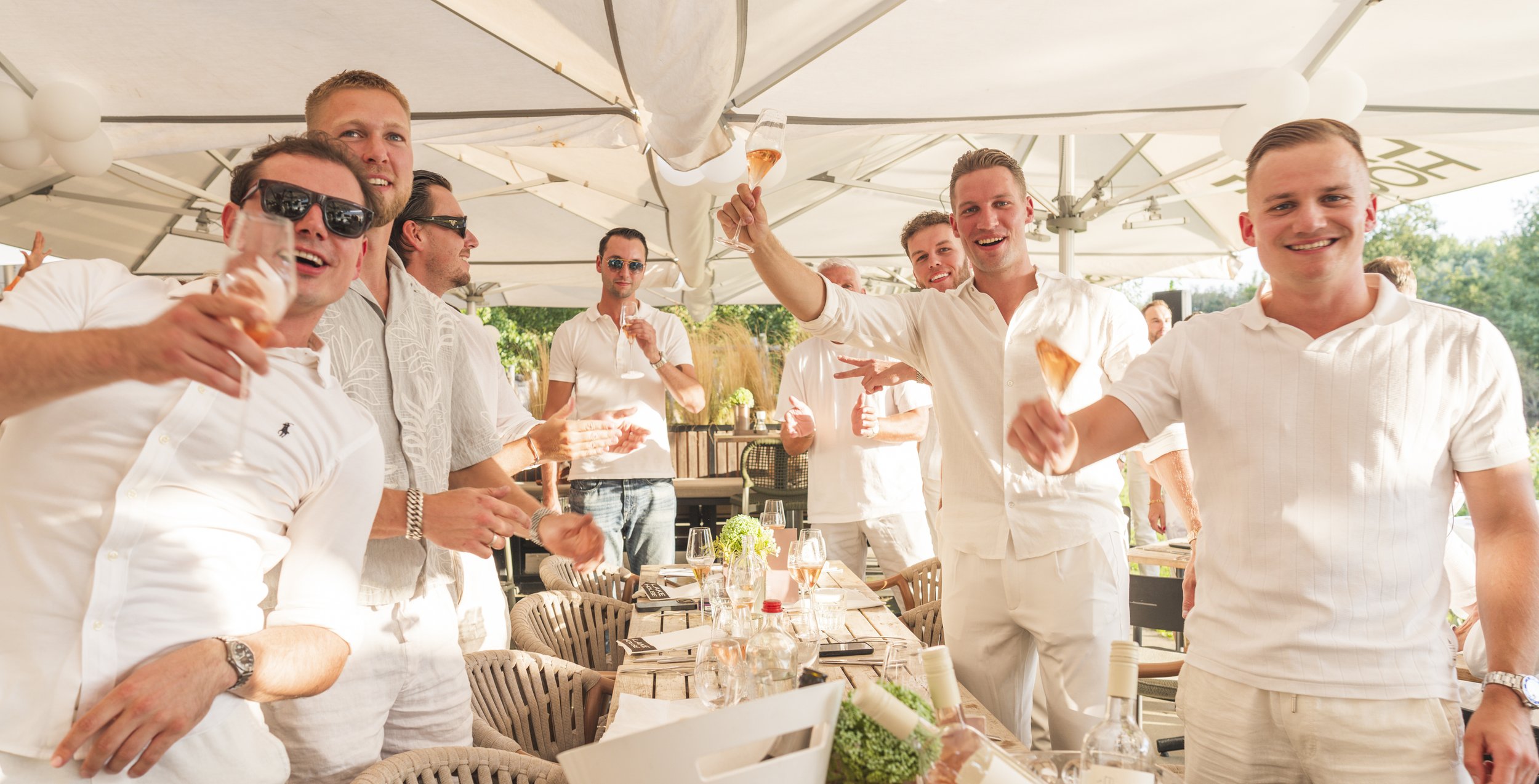 Groep mensen die samen viering of feestje bij een buitenrestaurant houden, met sommigen die wijn of champagne drinken en glimlachen.
