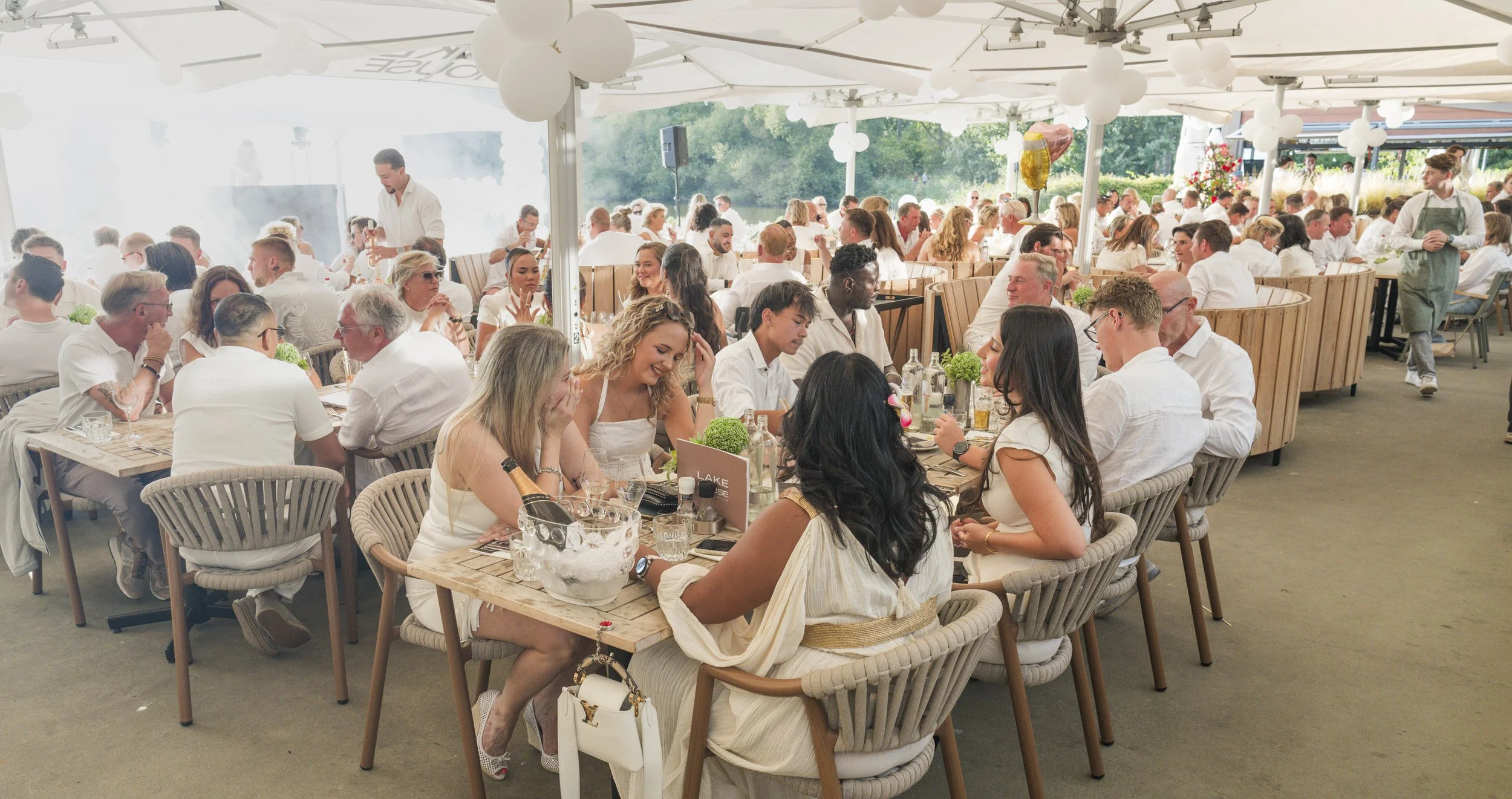 Een grote groep mensen zit samen aan lange houten tafels onder een witte tent met ballonnen en organiseert een feest of diner, het lijkt gezellig en alert.