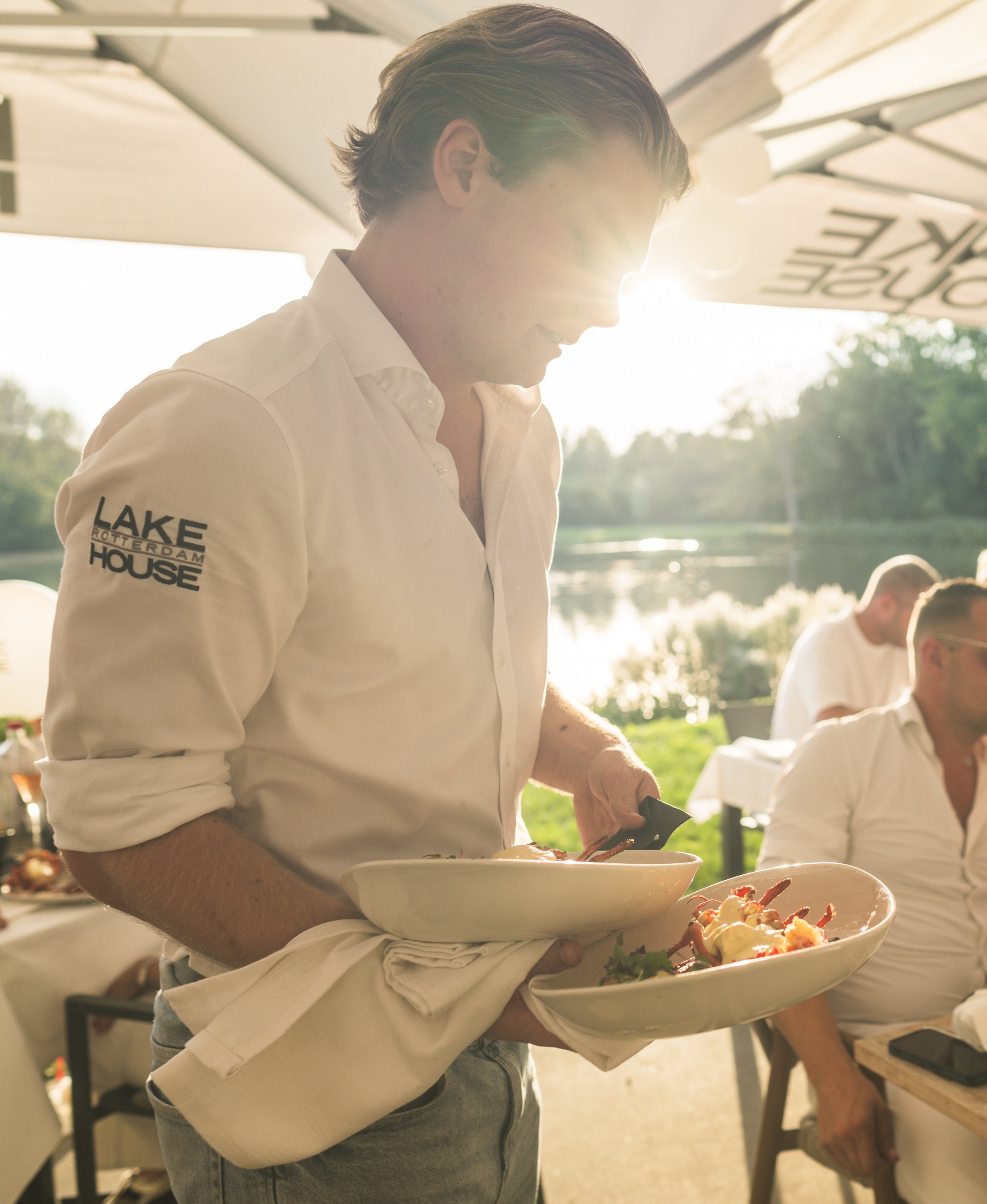Man houdt twee witte borden met eten, terwijl hij buiten staat met de zon op de achtergrond. Andere mensen zitten in de buurt.