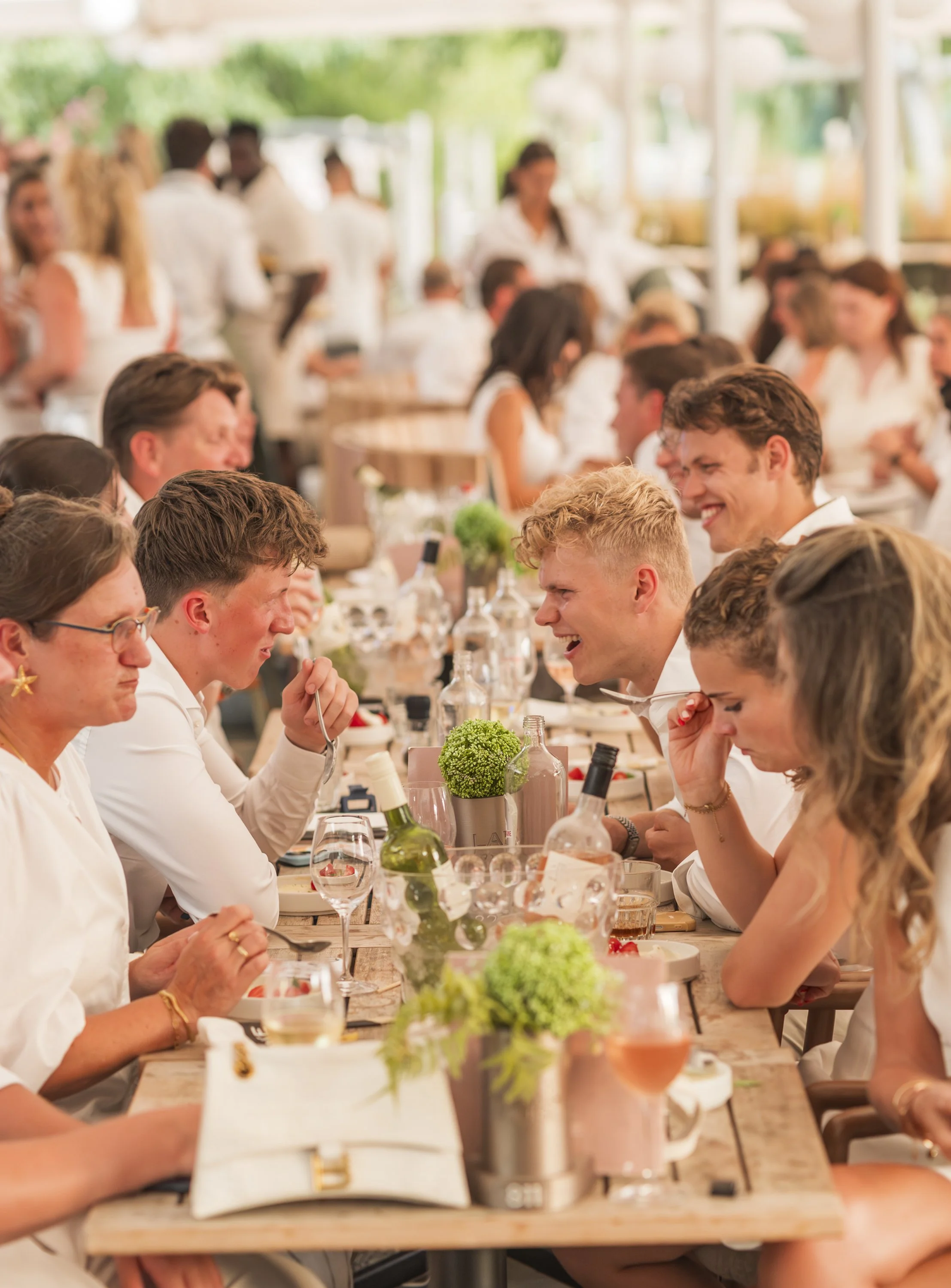 Groep mensen die aan een lange tafel buiten eten en lachen, met bloemen en wijn.