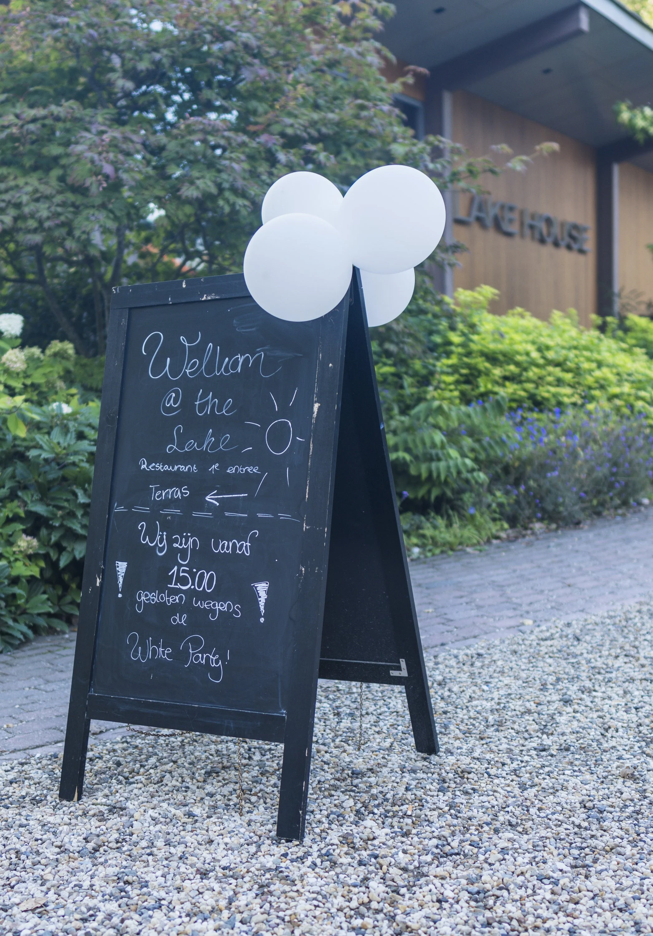 Slingerwhite bord met witte ballonnen en gedicht over White Party, buiten omgeving met tuin en gebouw in de achtergrond.