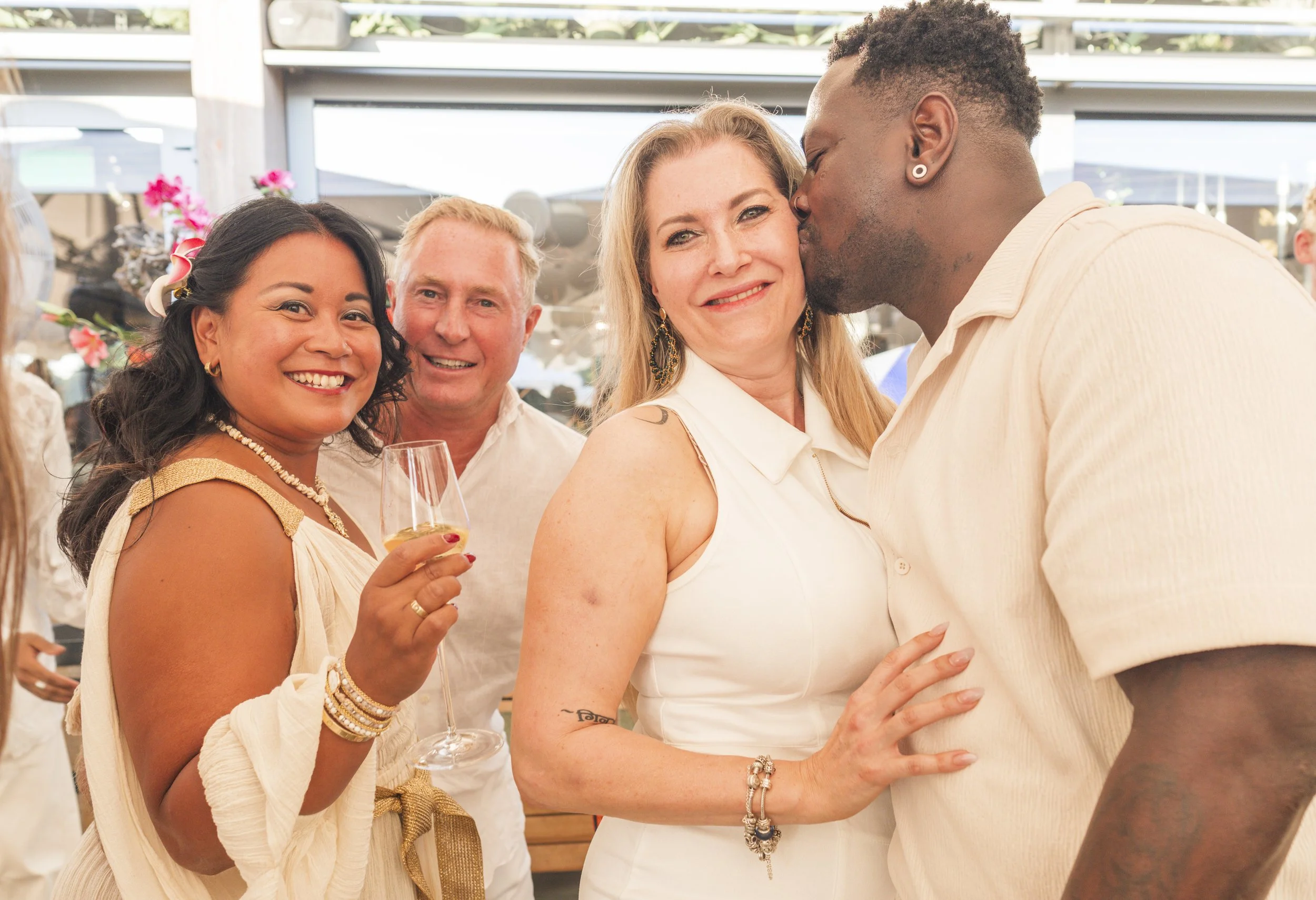 Vijf mensen die op een feestje genieten. Een vrouw houdt een glas wijn en glimlacht naar de camera, terwijl een man naast haar haar een kus geeft op het voorhoofd. Op de achtergrond zijn andere feestgangers en bloemstukken te zien.