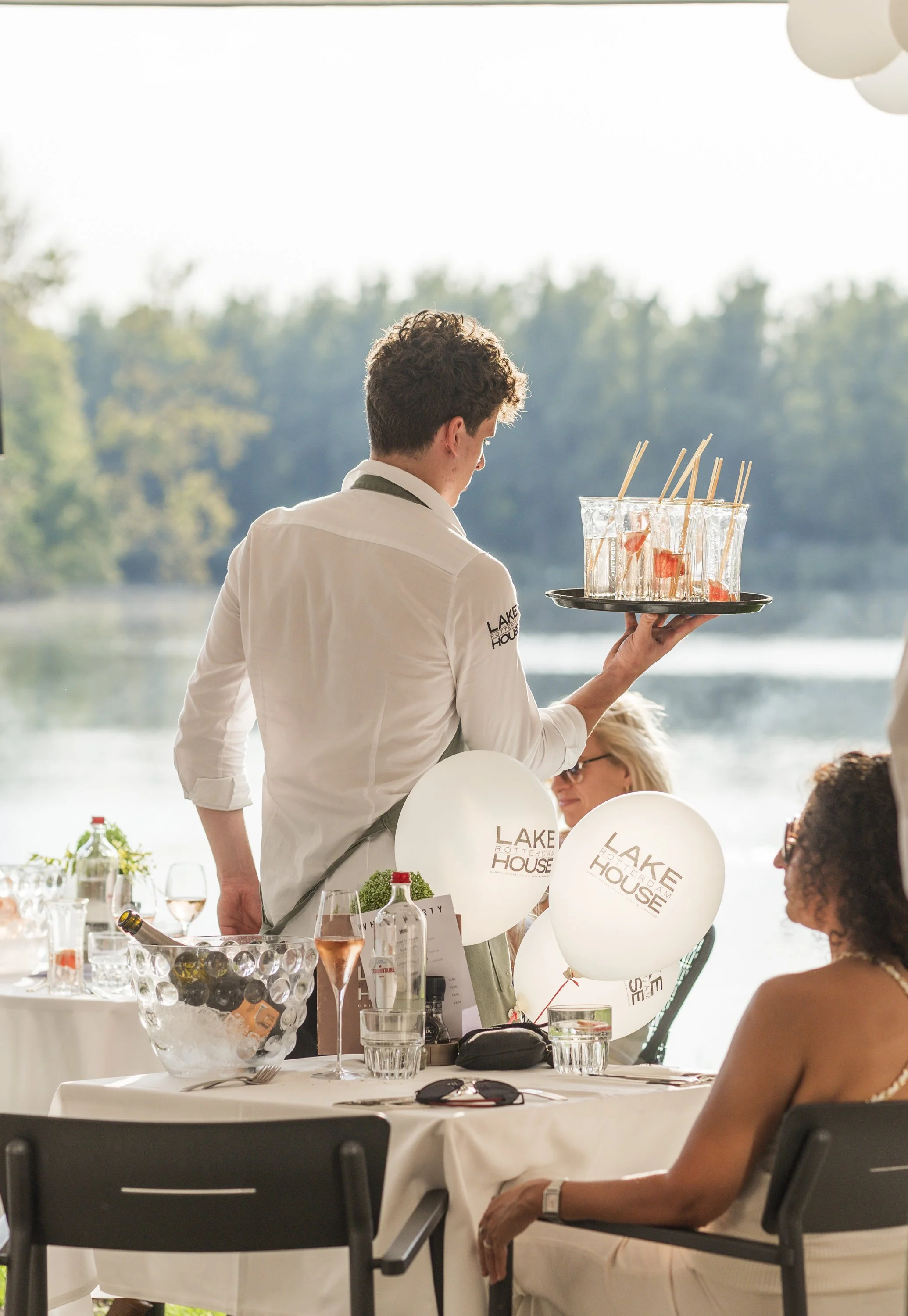 Een man in een witte blouse serveert drankjes met rietjes op een feest of diner bij een tafel met glazen, een fruitschaal en ballonnen met de tekst 'Lake House'.