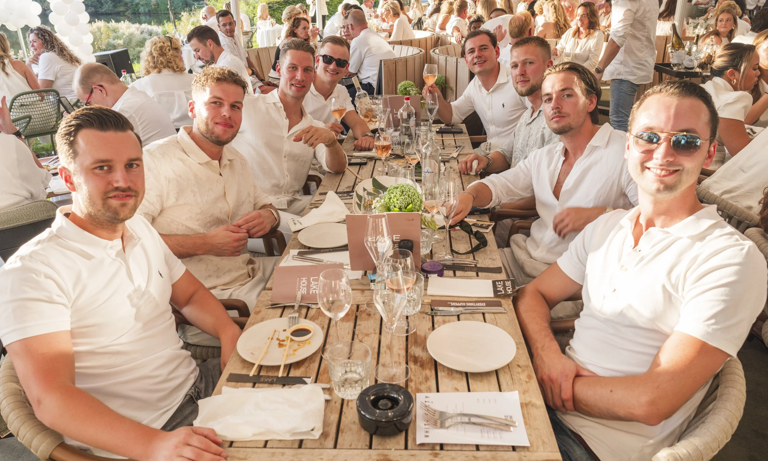 Een groep jonge mannen die aan een lang houten tafel zitten tijdens een buitenevenement, met sommigen die wijn drinken en etensergen op tafel, terwijl een levendige feestelijke sfeer wordt weergegeven.