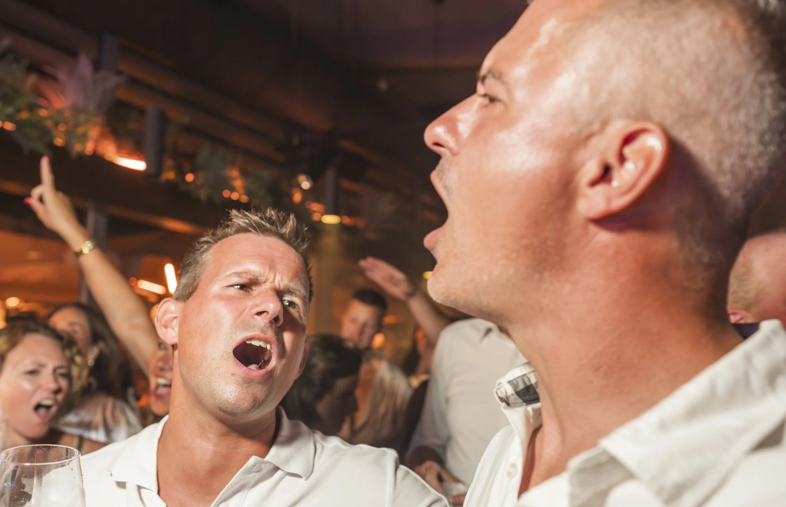Groep mensen die op een feestje plezier hebben, praten en lachen in een drukke, gezellige setting met warme verlichting.