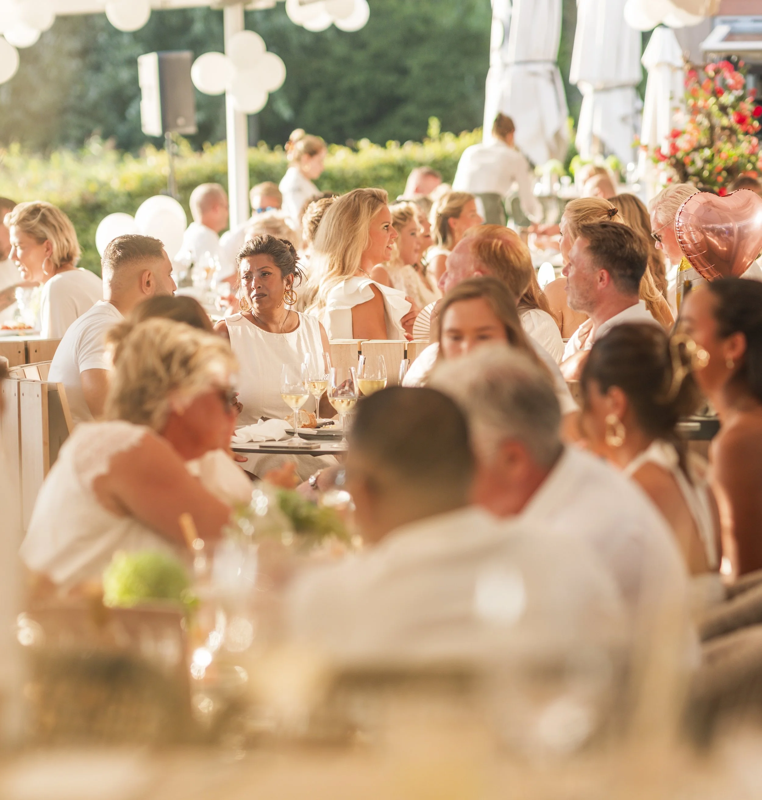 Een feestelijke setting met mensen die aan lange tafels zitten, lachend en pratend, onder een overkapping met decoraties en ballonnen, buiten in de zon.