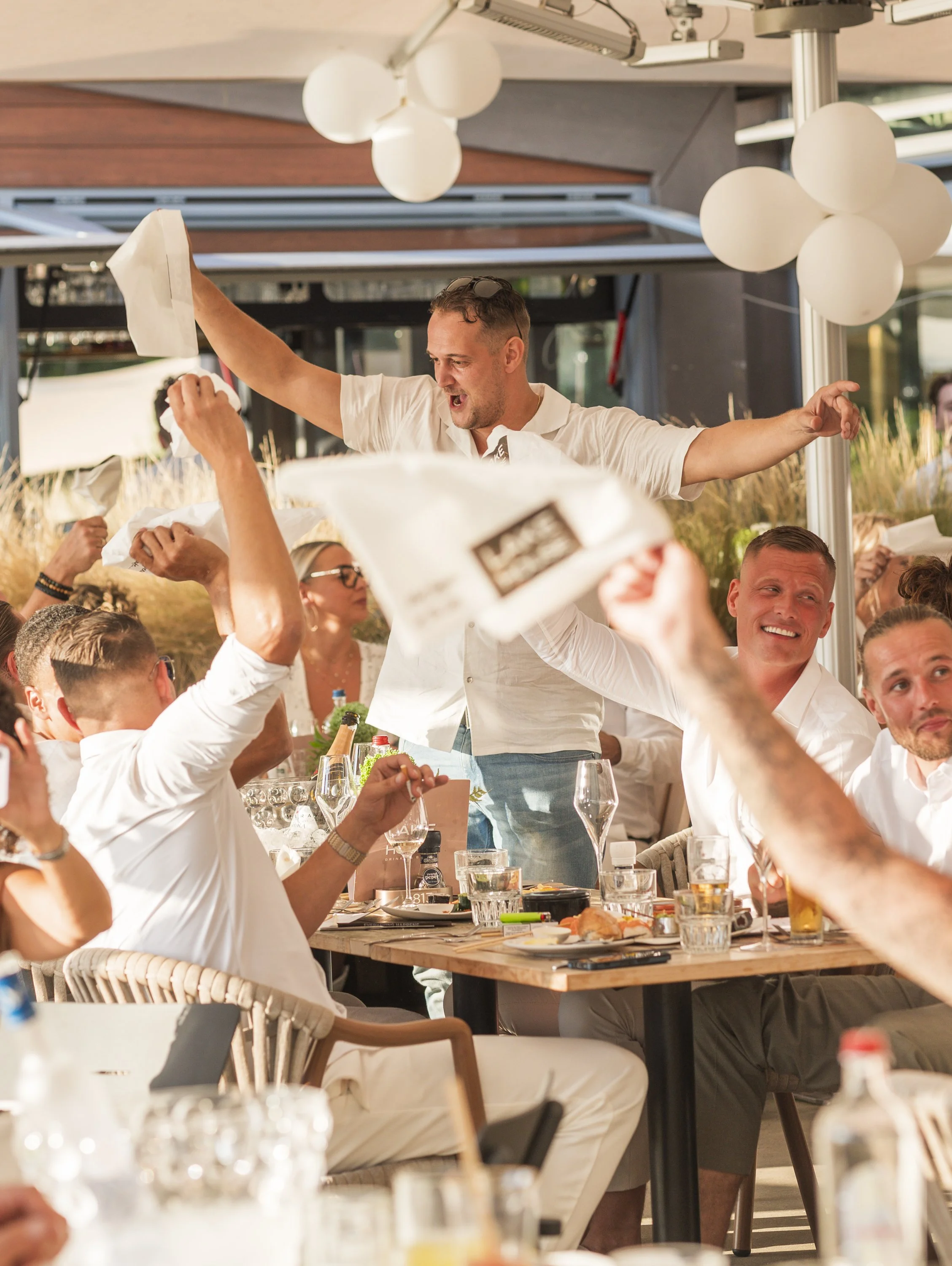 Manen en vrouwen aan een feesttafel met eten en drinken, mensen die juichen en een man die feestelijk springt op een stoel, decoraties met witte ballonnen, buiten op een terras.