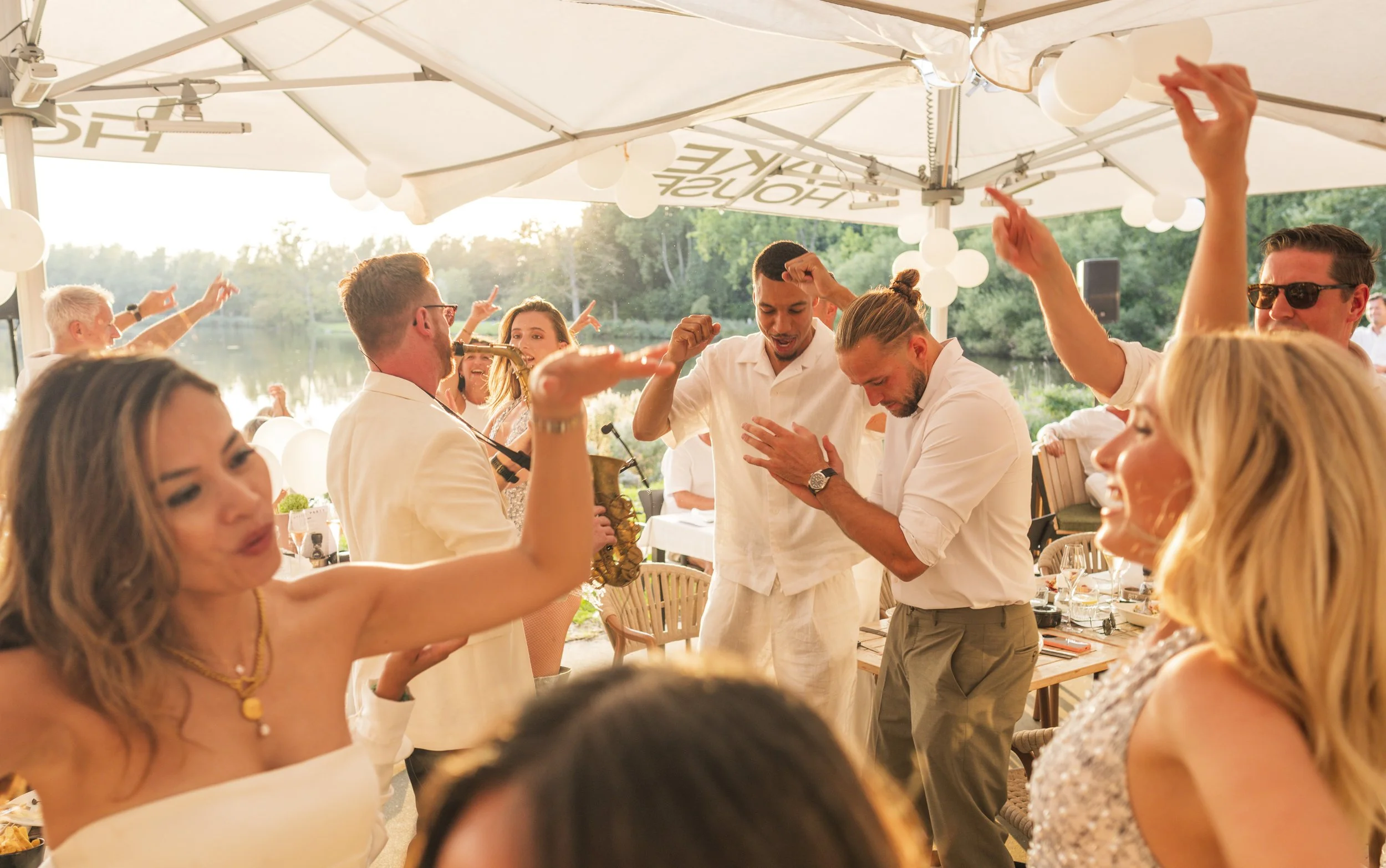 Gezelschap mensen die dansen en genieten op een feestje onder een grote parasol bij een waterpartij, omgeven door groen natuur.