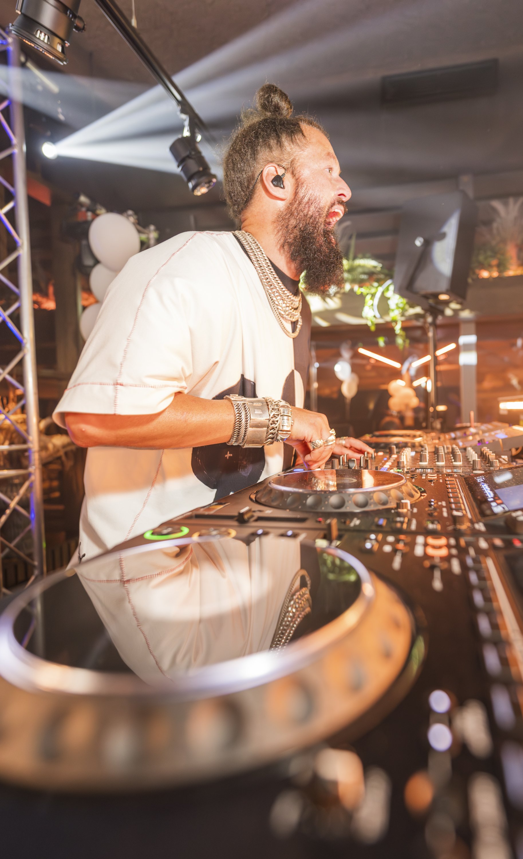 De afbeelding toont een man DJ met een lange baard en een man bun, die op een ergonomisch DJ-pult staat en aan het draaien is op een feest of club, met kleurrijke verlichting en geluidssystemen.