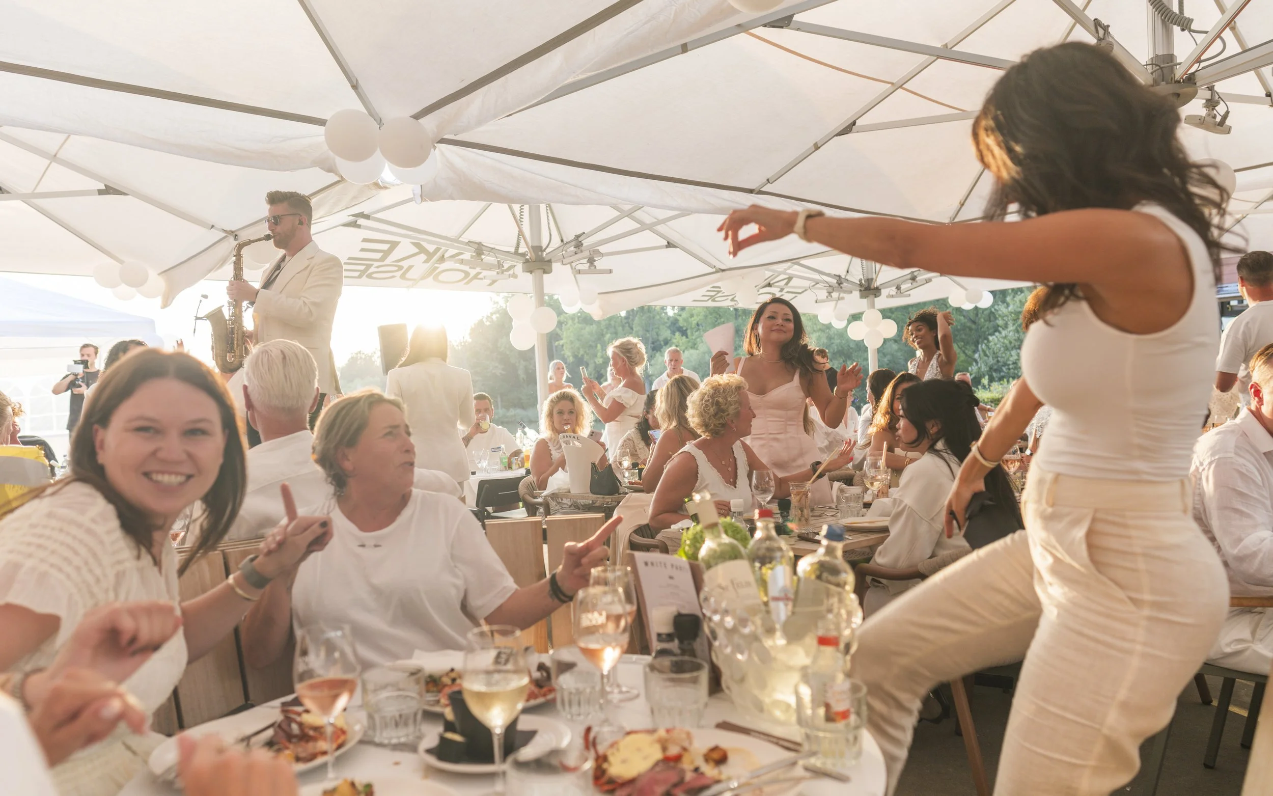 Mensen genieten van een feest onder een grote witte parasol, met een muzikant die saxofon speelt op de achtergrond, terwijl ze dansen en eten.