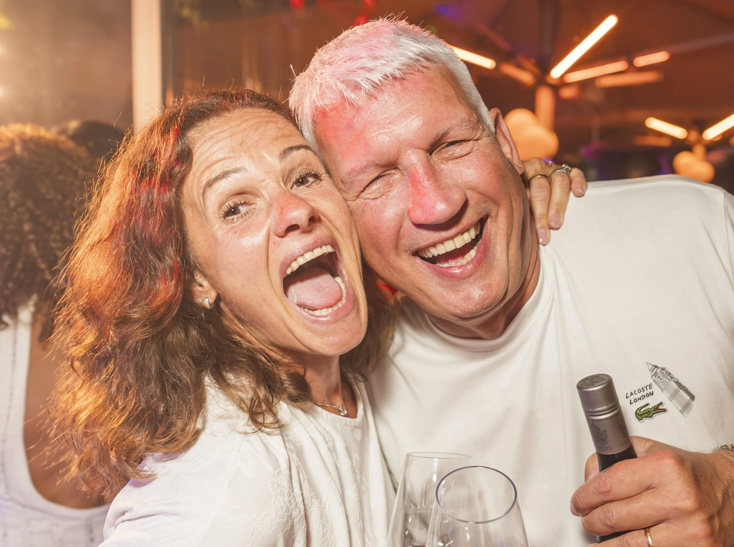 Twee mensen lachen en genieten, één vrouw met rood haar en één man met wit haar, in een feestsetting met kleurrijk licht, ze houden champagneglazen vast en de man gebruikt een lippenstift.