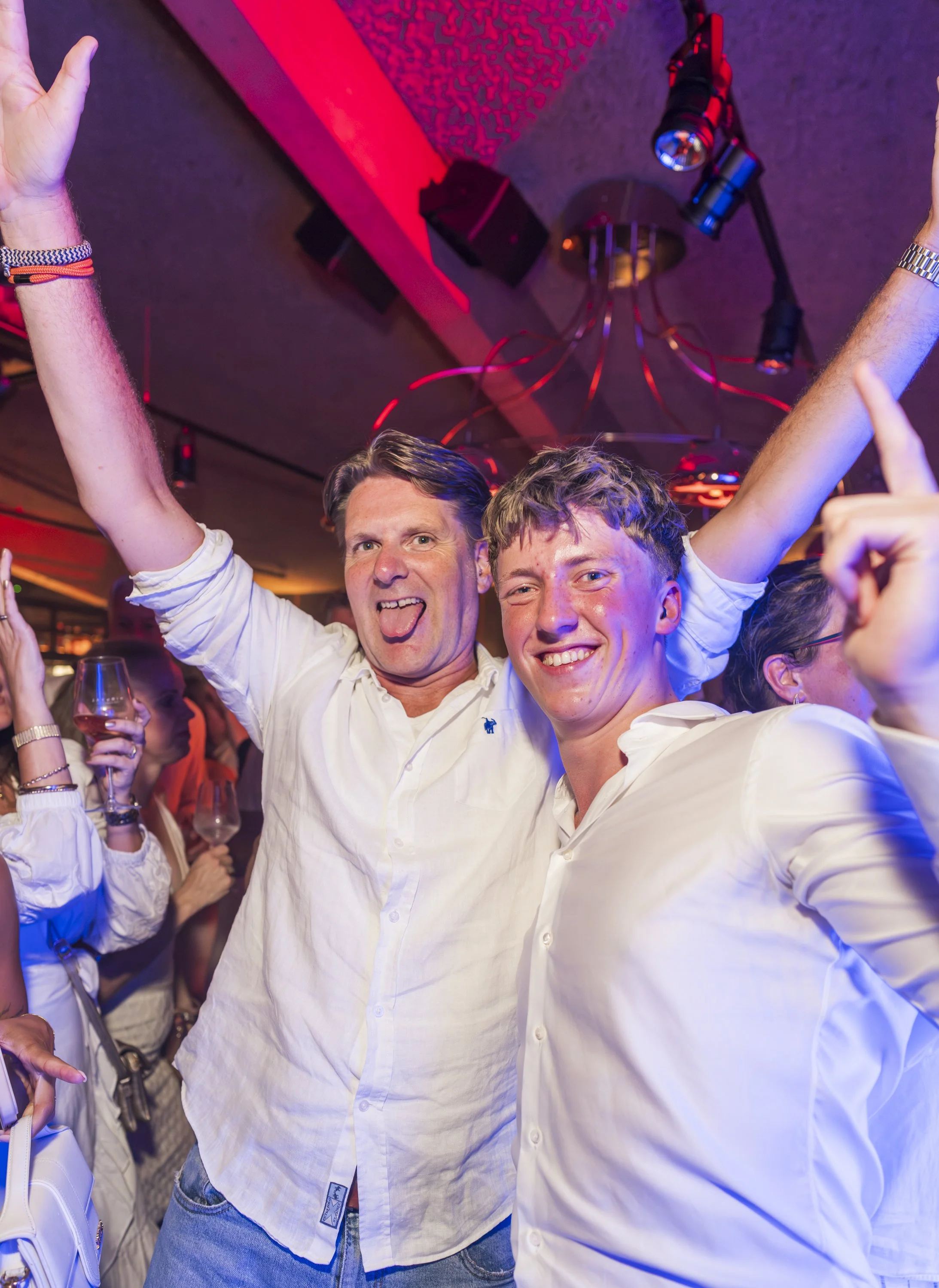 Twee jonge mannen glimlachen en gooien hun handen in de lucht tijdens een gezellig feestje, met kleurrijke verlichting en andere feestgangers op de achtergrond.