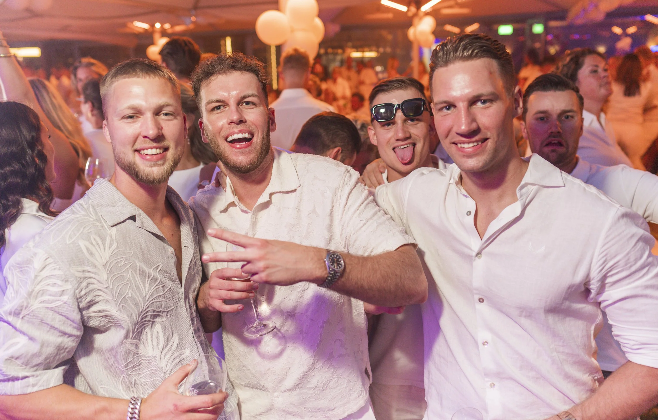 Vijf jonge mannen die plezier maken op een feestje, lachen, poseren voor de camera, met een glas wijn in een drukke, kleurrijke feestzaal.