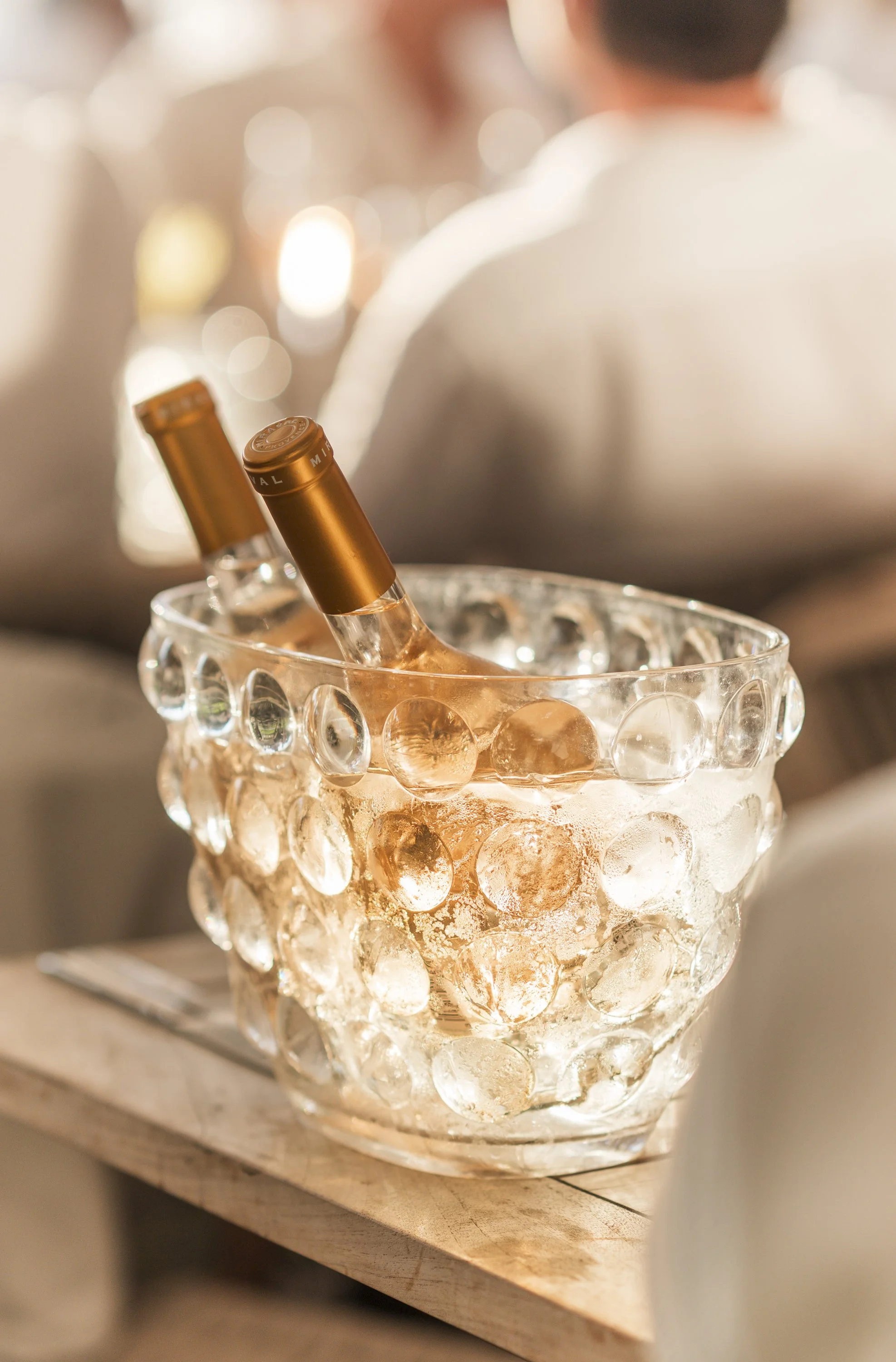 Een glazen kom met lichtgevend ijsblokjes en twee oplichtende champagneflessen binnenin, geplaatst op een houten ondergrond.